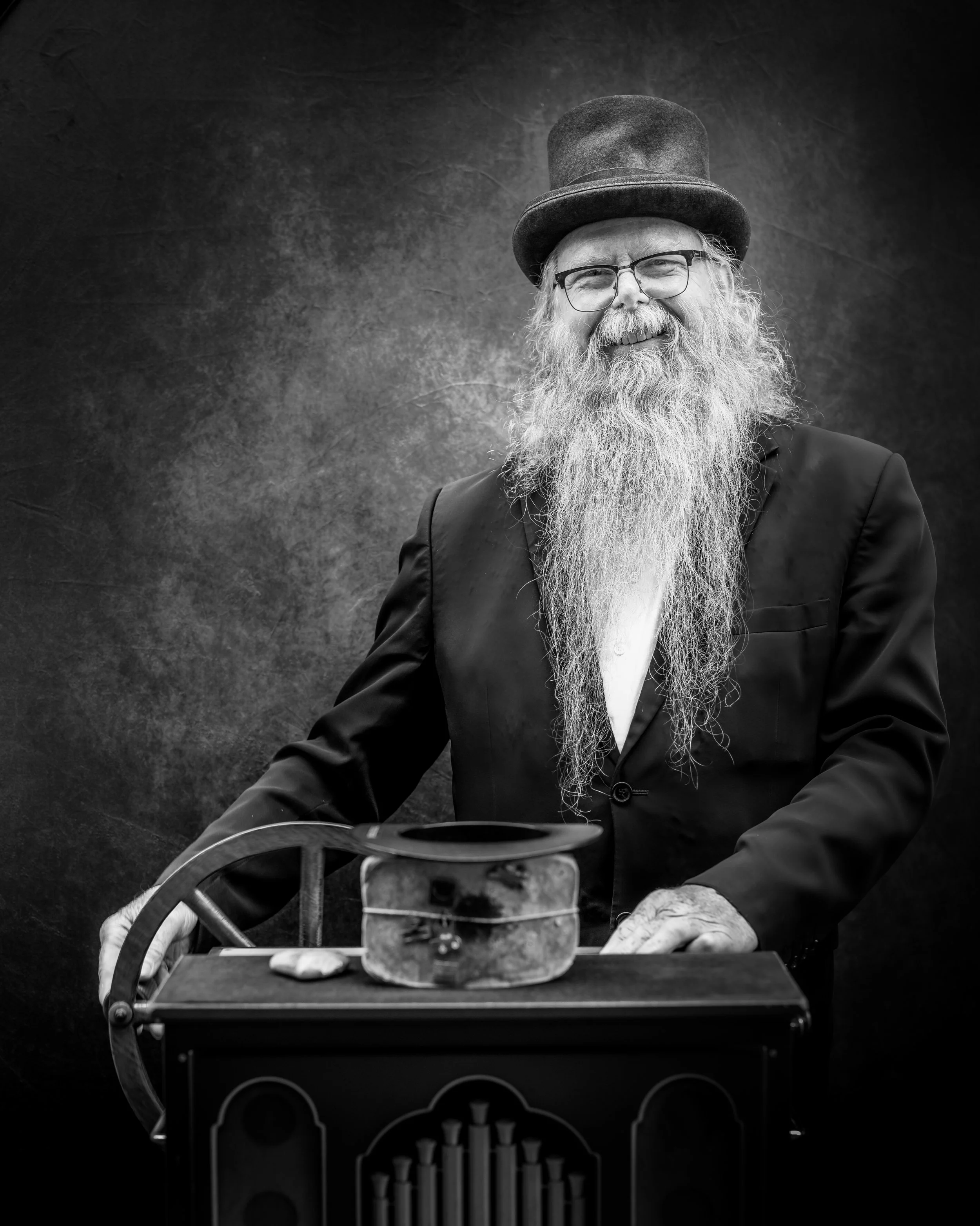 A smiling elderly man with a long white beard, wearing glasses, a top hat, and a suit, standing behind a vintage musical instrument in a studio with a dark textured background.