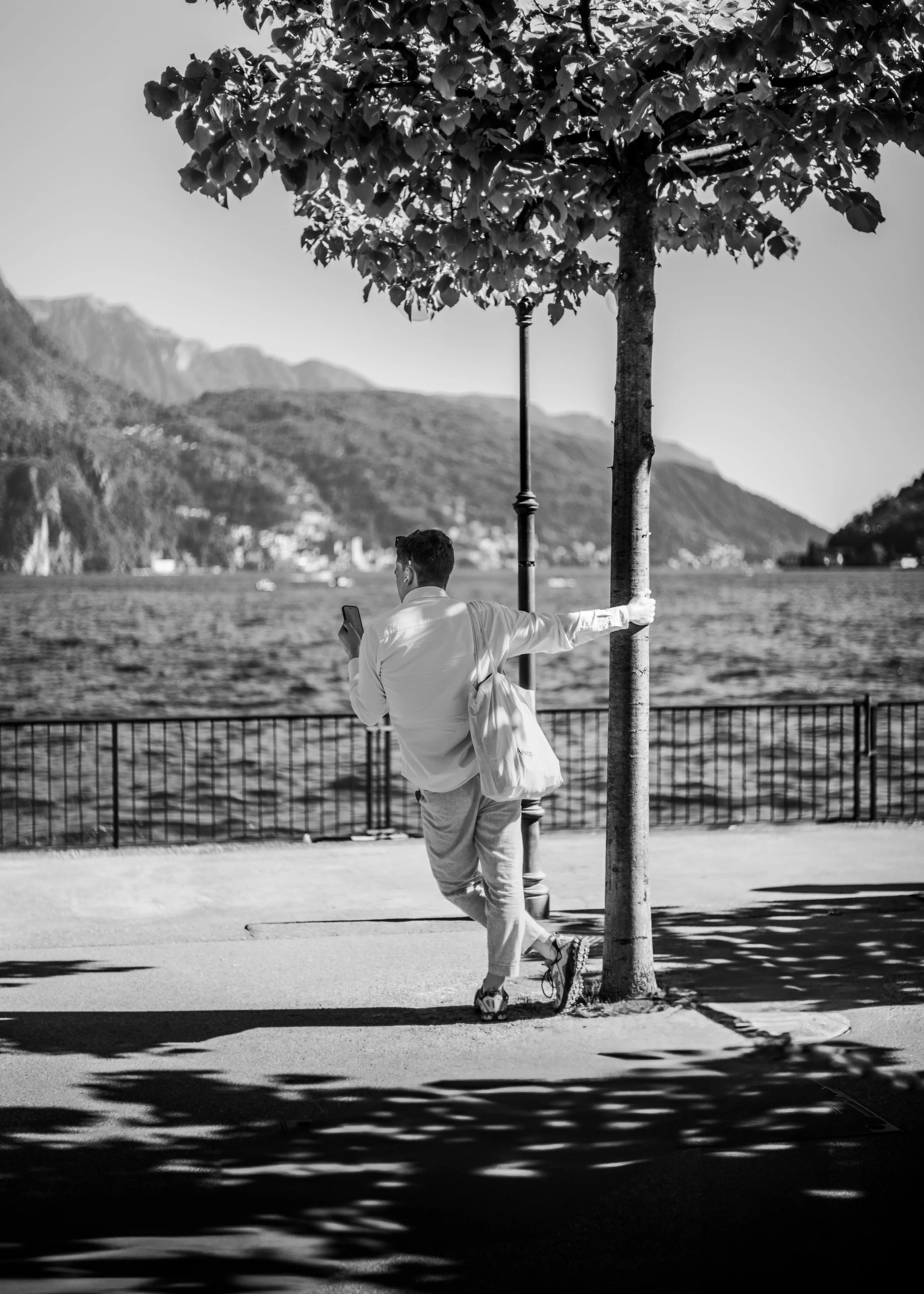 A man in light-colored pants and a long-sleeve shirt leaning against a tree while looking at his phone near a waterfront with mountains in the background, in black and white.