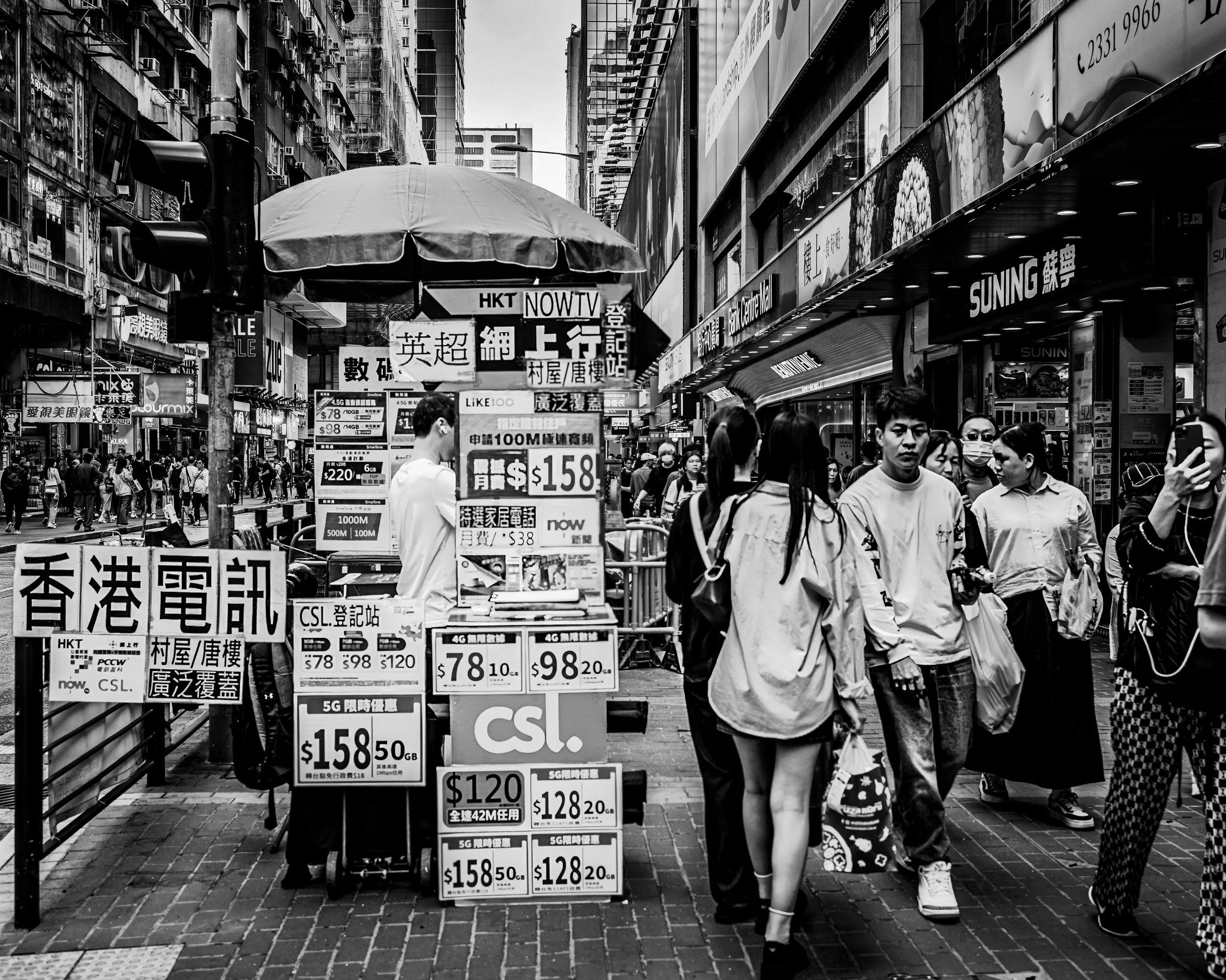 A busy urban street scene with pedestrians walking past storefronts. There are signs and advertisements in Chinese and English, including a street vendor selling SIM cards with various prices. The scene is in black and white.