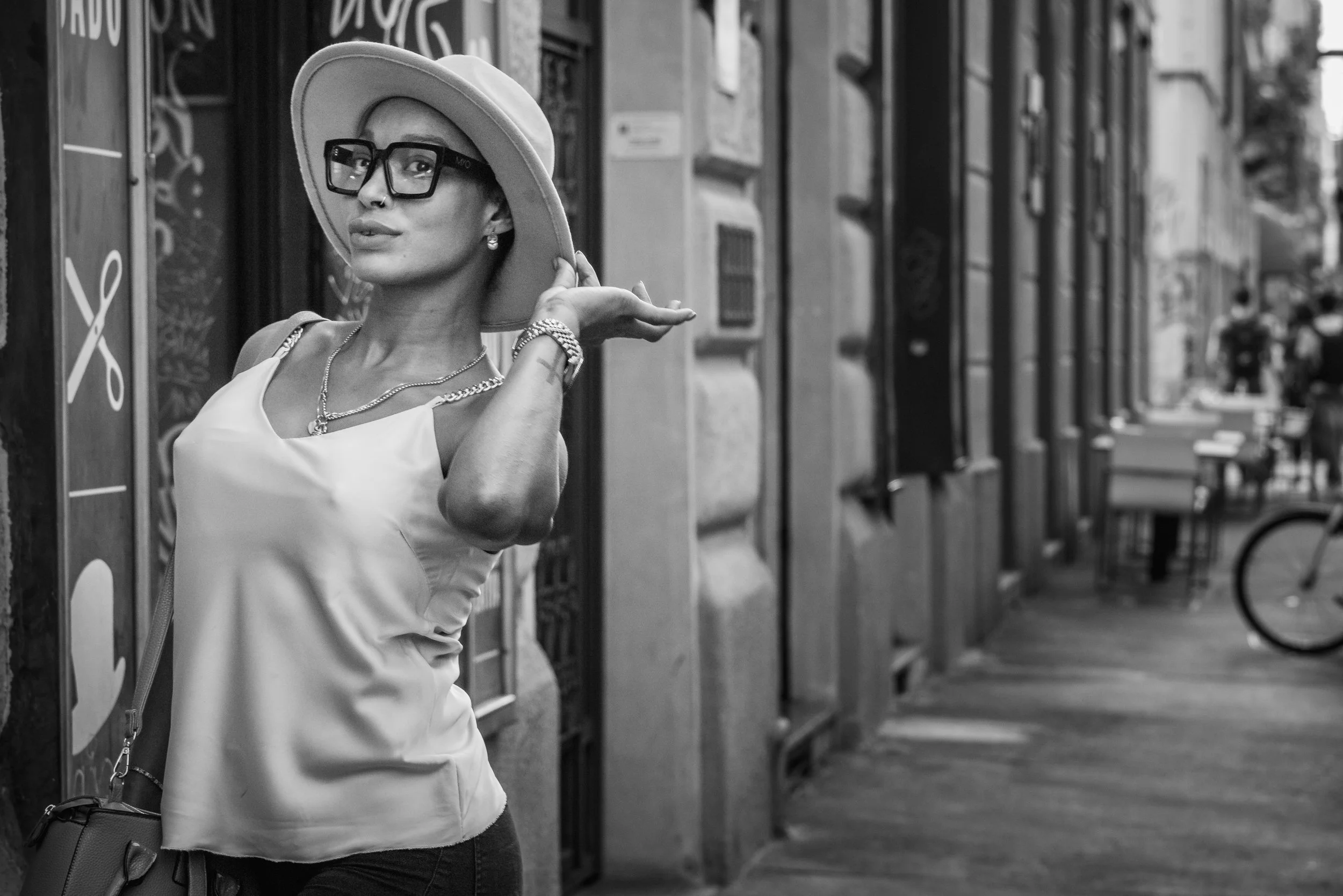 A woman with short hair wearing a wide-brimmed hat, large sunglasses, jewelry, and a satin sleeveless top posing on a city sidewalk.