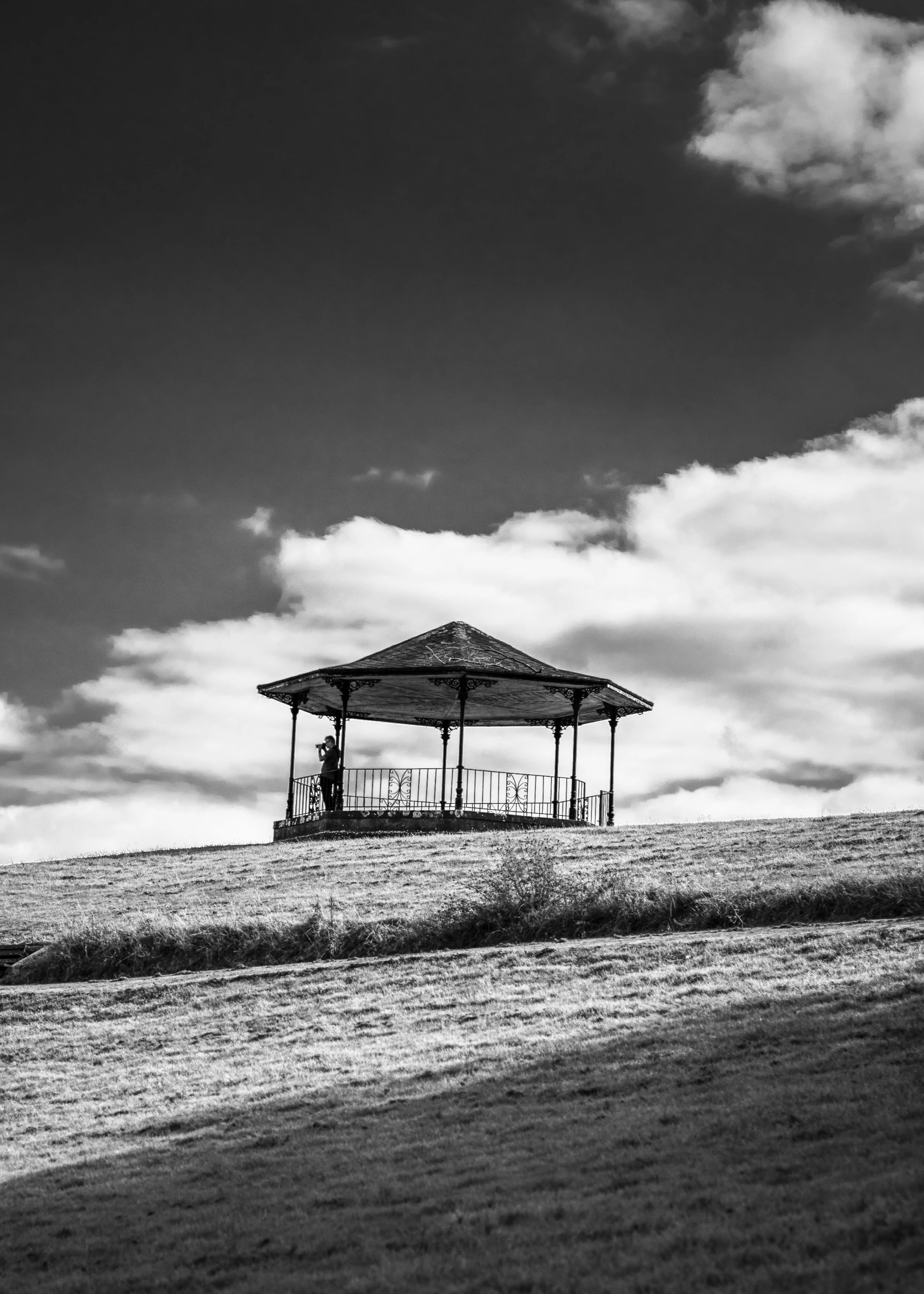A black and white photo of a gazebo on a grassy hill with a person standing inside, looking through binoculars, under a cloudy sky.
