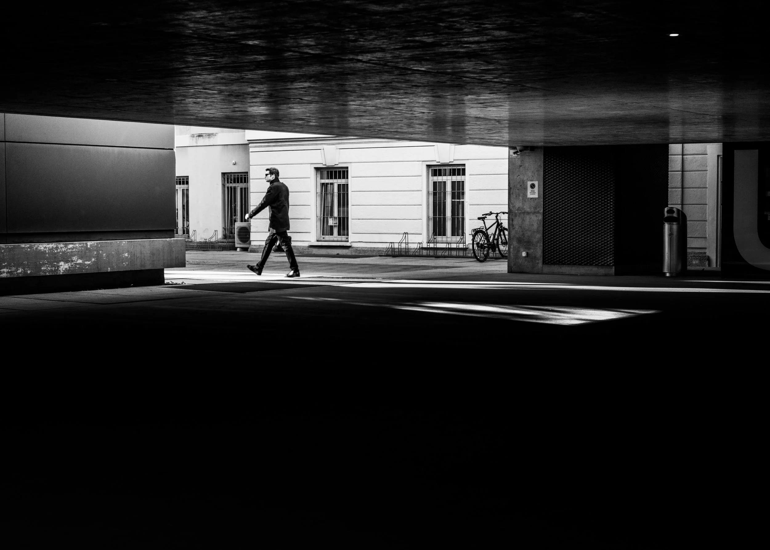 A man walks across a street beneath a bridge or overpass in a city, with bicycles parked against a building wall and a trash bin nearby, in black and white.