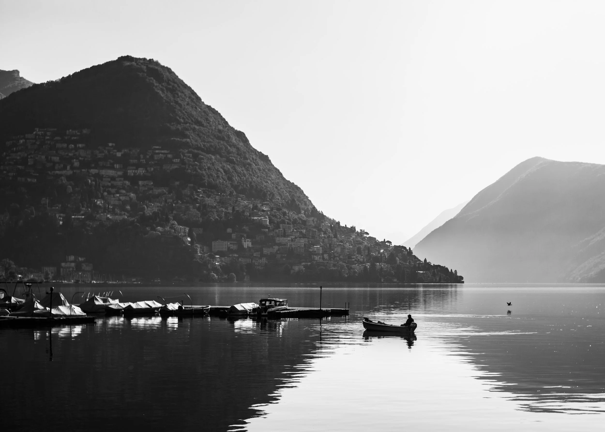 A black and white photo of a lake with boats, surrounded by hills or mountains with houses along the shoreline.