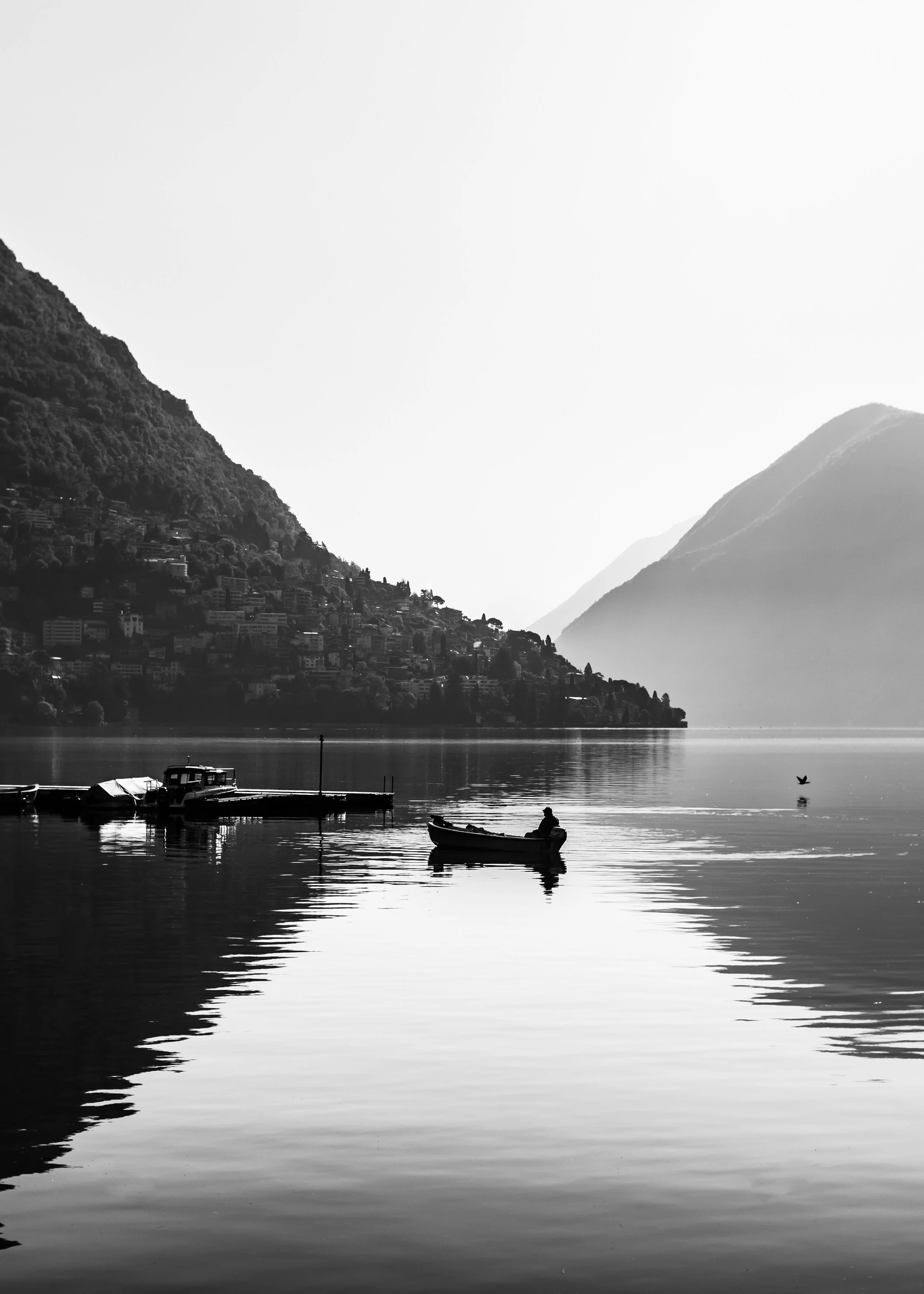 Black and white photo of a lake with boats, surrounded by mountains and a small hillside town in the background.