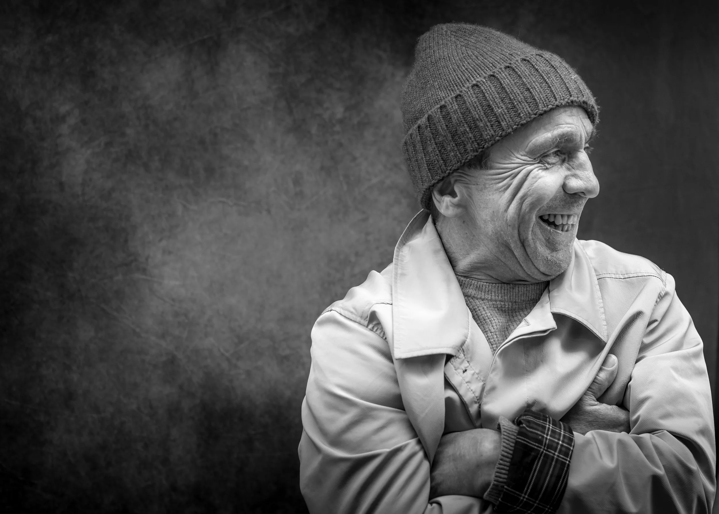 Black and white photo of an elderly man laughing, wearing a knit beanie and jacket, with a dark background.