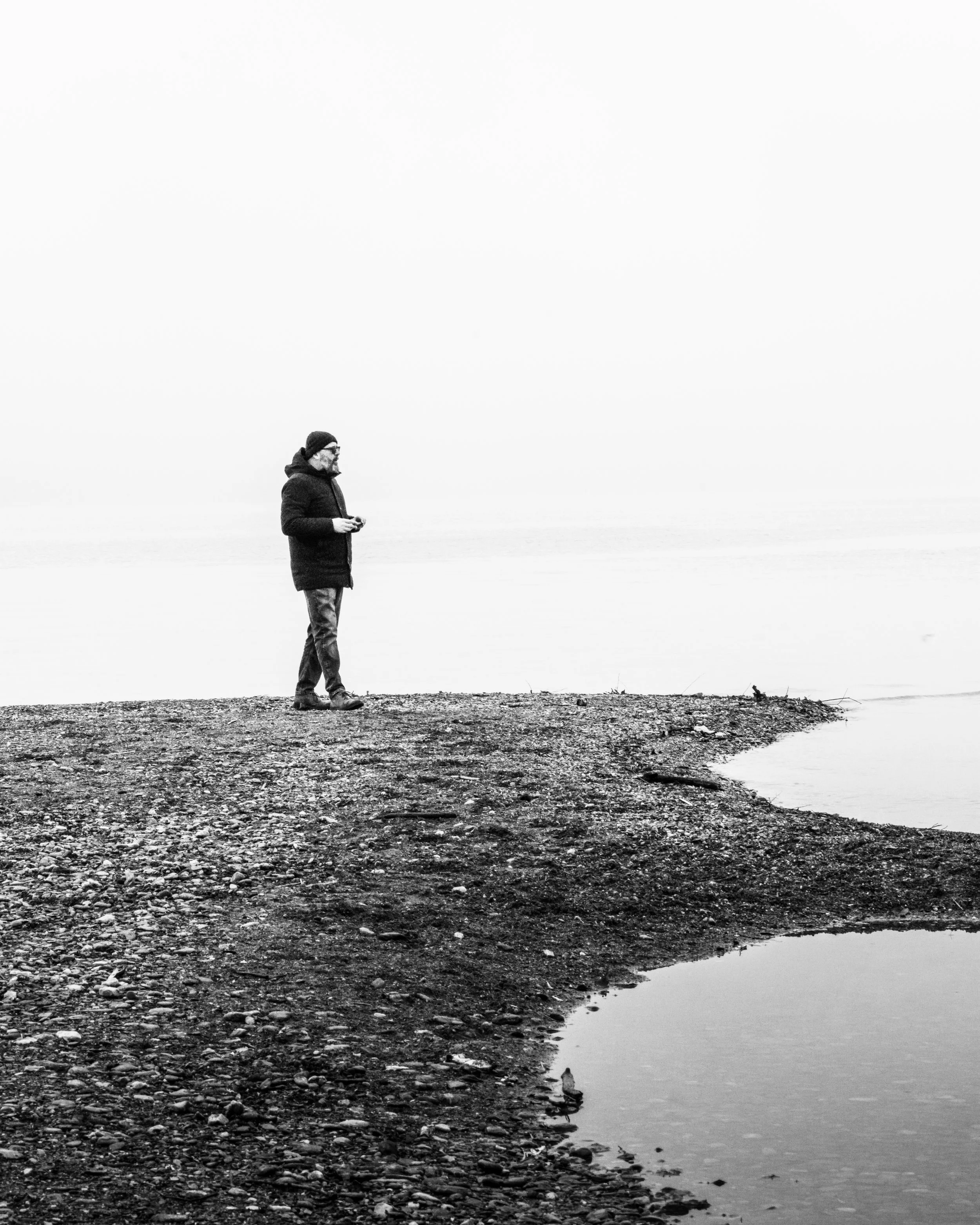 A man in winter clothing standing on a rocky shoreline looking towards water with a foggy or overcast background in black and white.