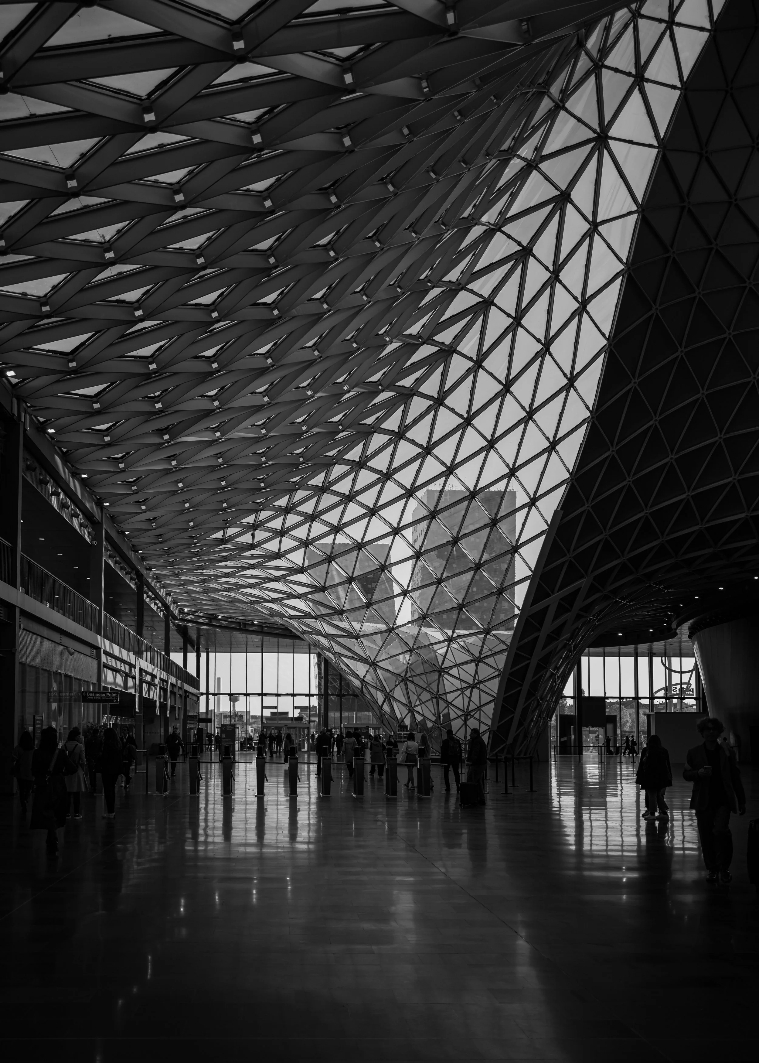 Interior of a modern building with a curved, glass and steel roof structure, with people walking and standing inside.