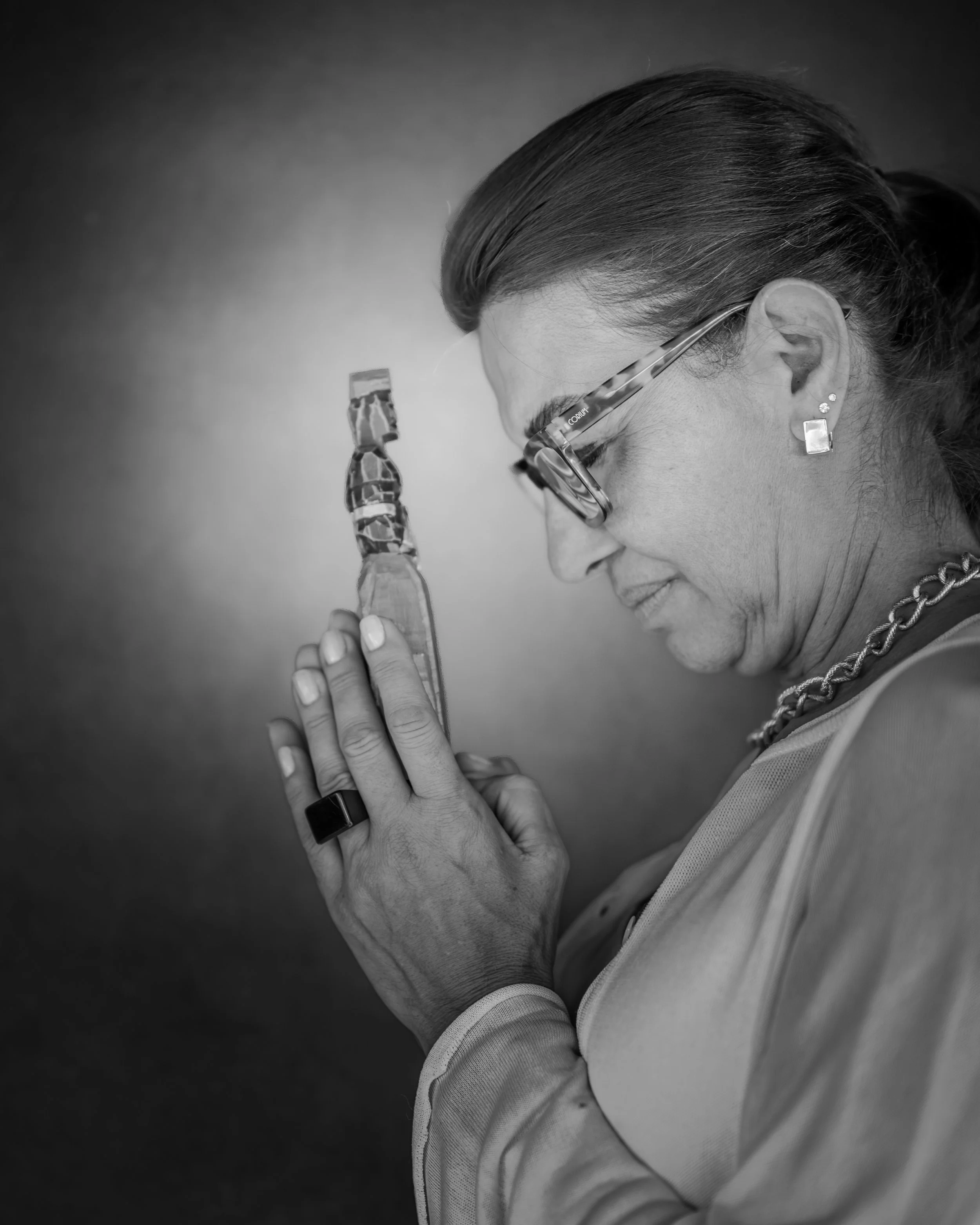 Black and white photo of a woman with glasses and earrings holding her hands in a prayer position, with a bottle pressed between her palms, eyes closed, and her head bowed.