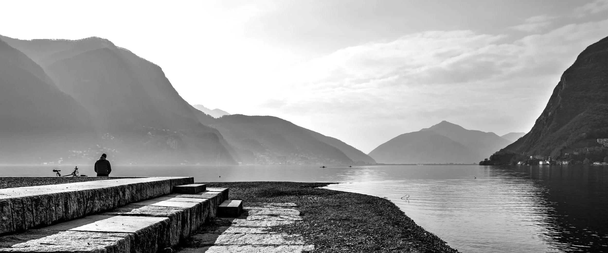A black and white photo of a person sitting on a stone pier facing a calm lake surrounded by mountains, with a bicycle beside them.