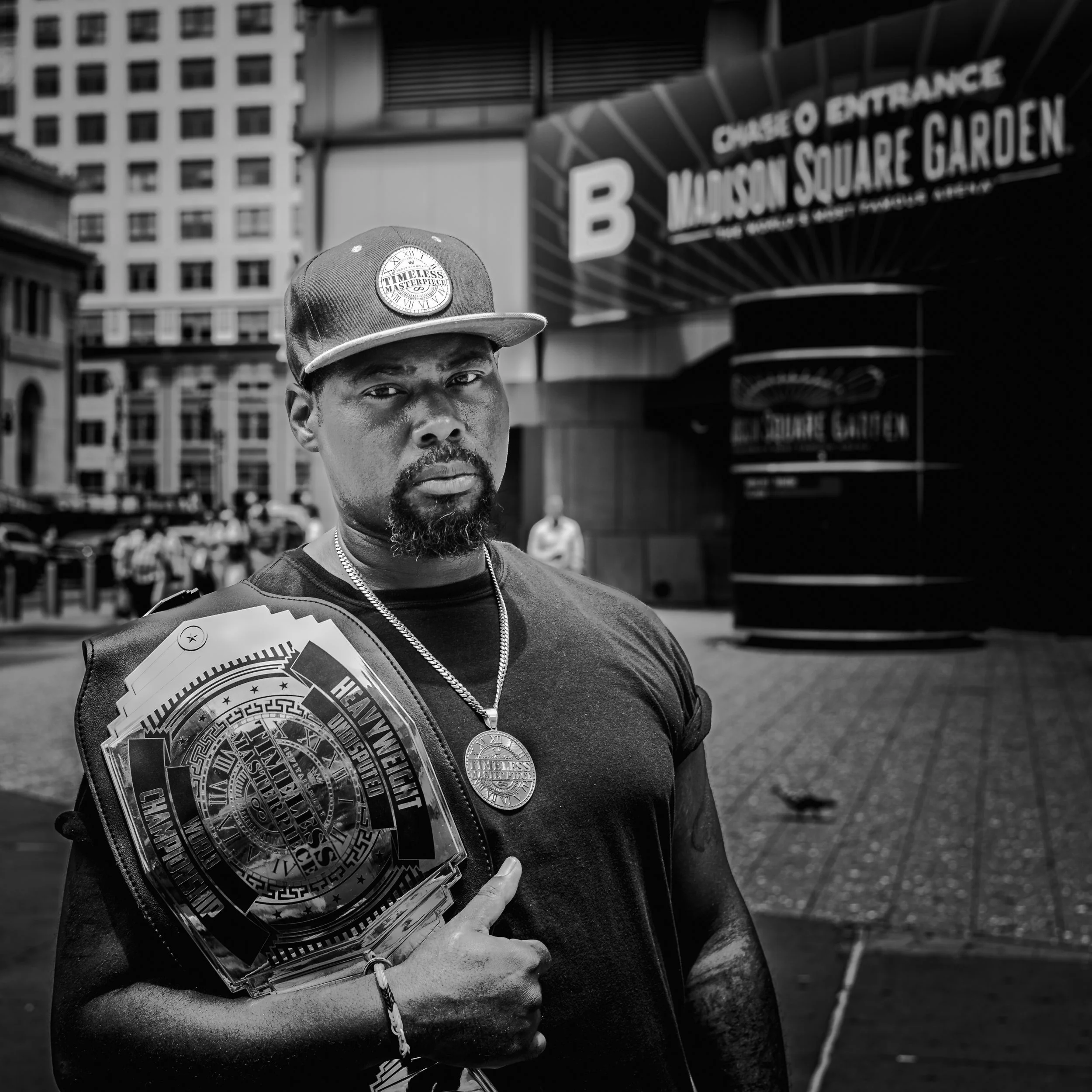 A man wearing a cap, black t-shirt, and medal around his neck, standing outside Madison Square Garden with championship belts over his shoulder, giving a thumbs-up.