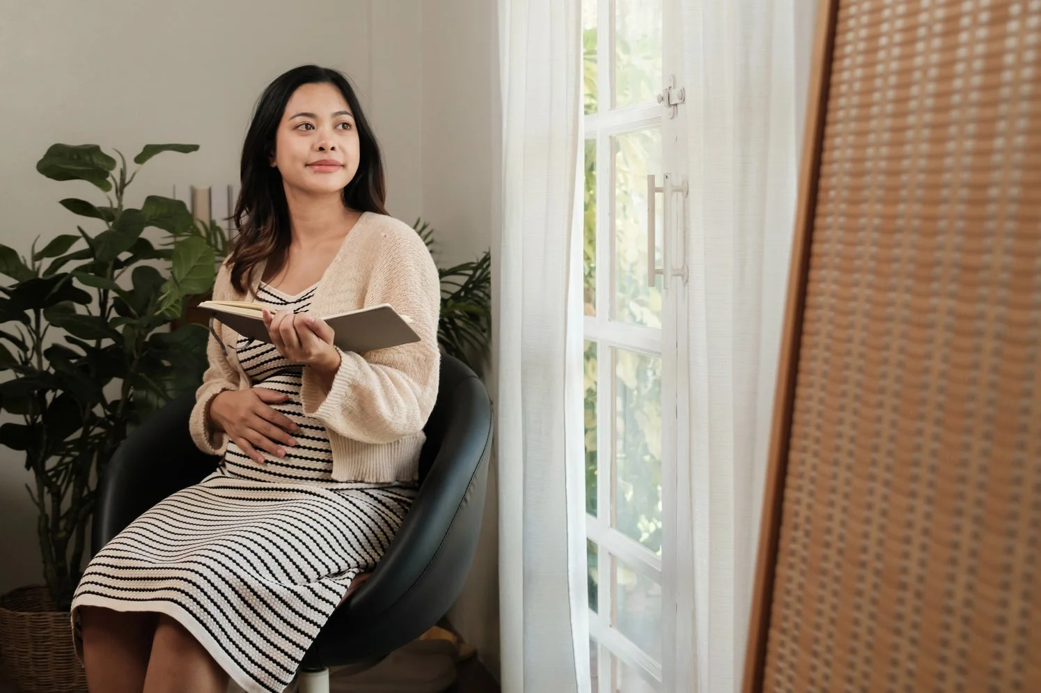 A pregnant Asian woman sitting in a black armchair, cradling her bump with one hand while holding an open journal and looking out a window.