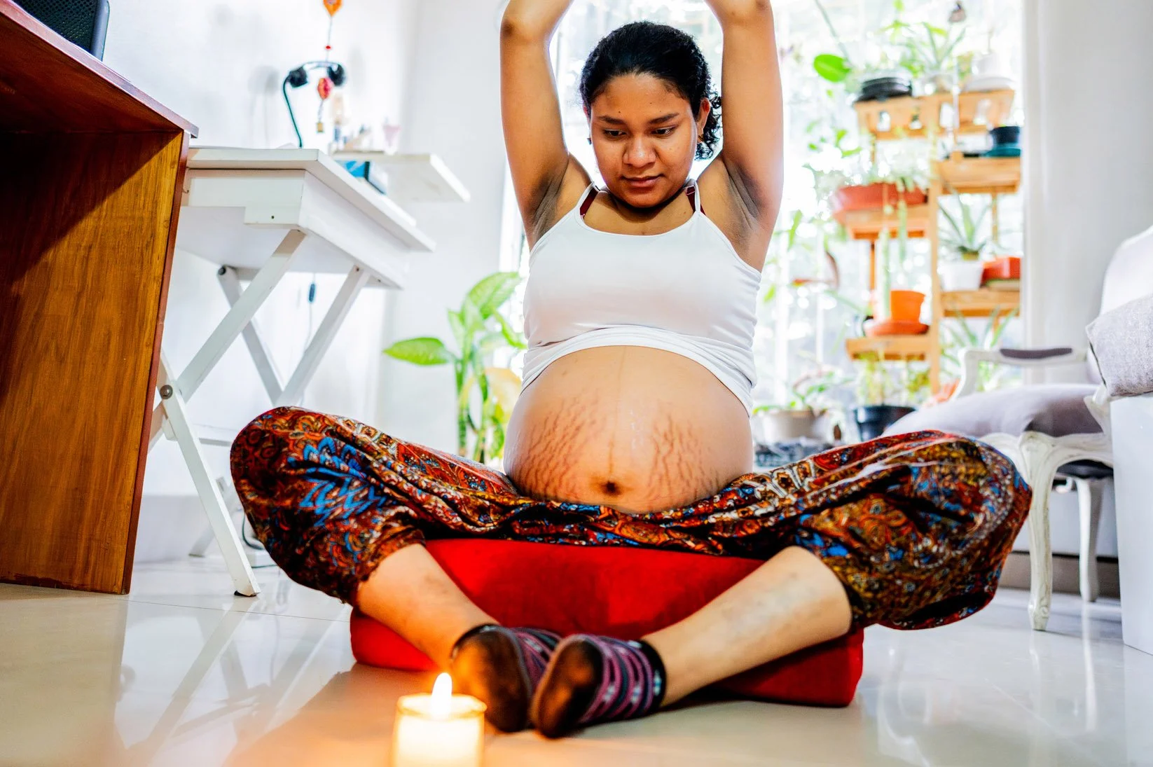 A pregnant woman sitting cross-legged on a floor cushion with her arms raised overhead in a yoga stretch, looking down at a lit candle.