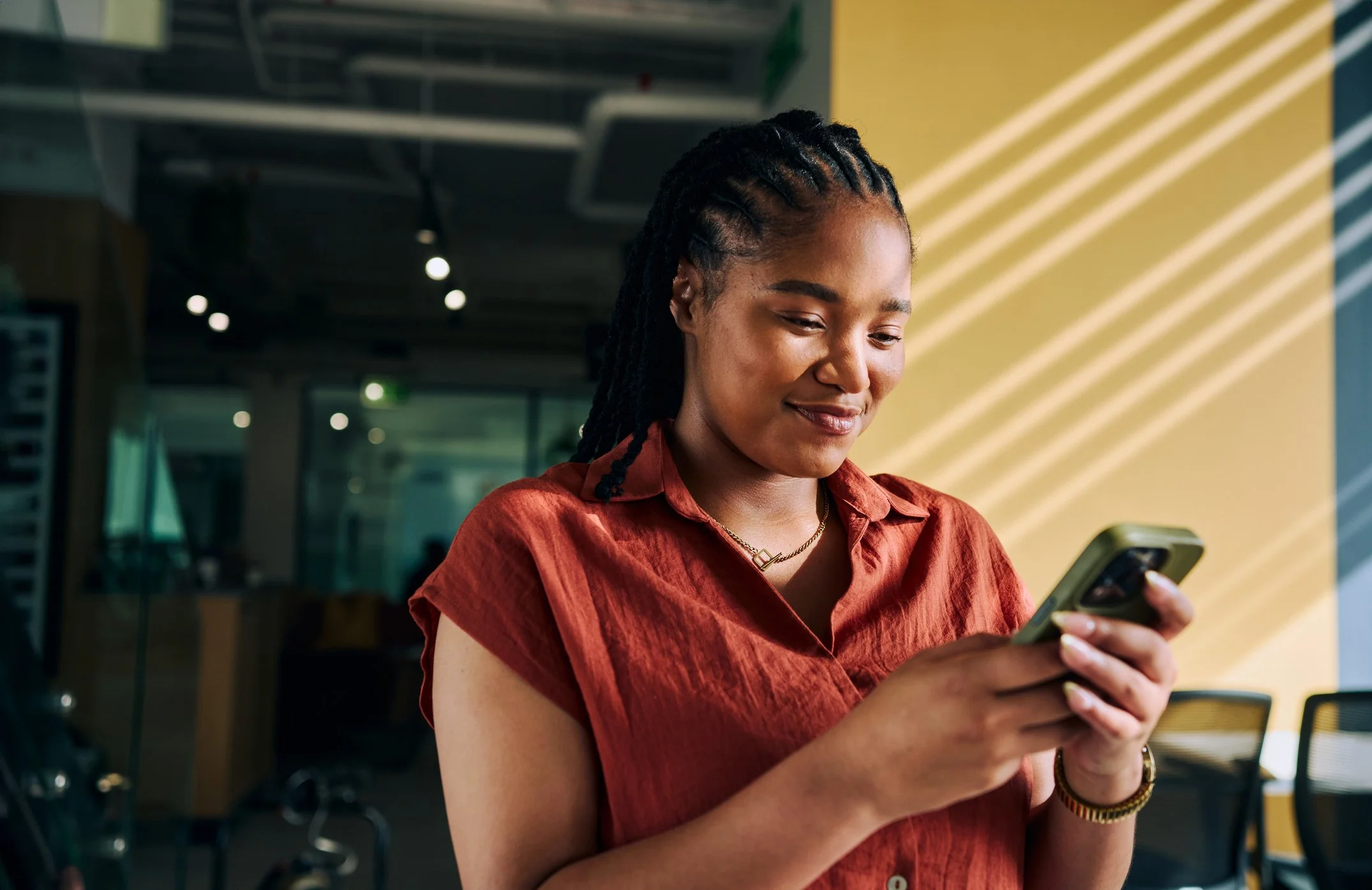A Black woman with braided hair wearing a rust-red shirt, smiling gently down at her smartphone in a sunlit modern office.