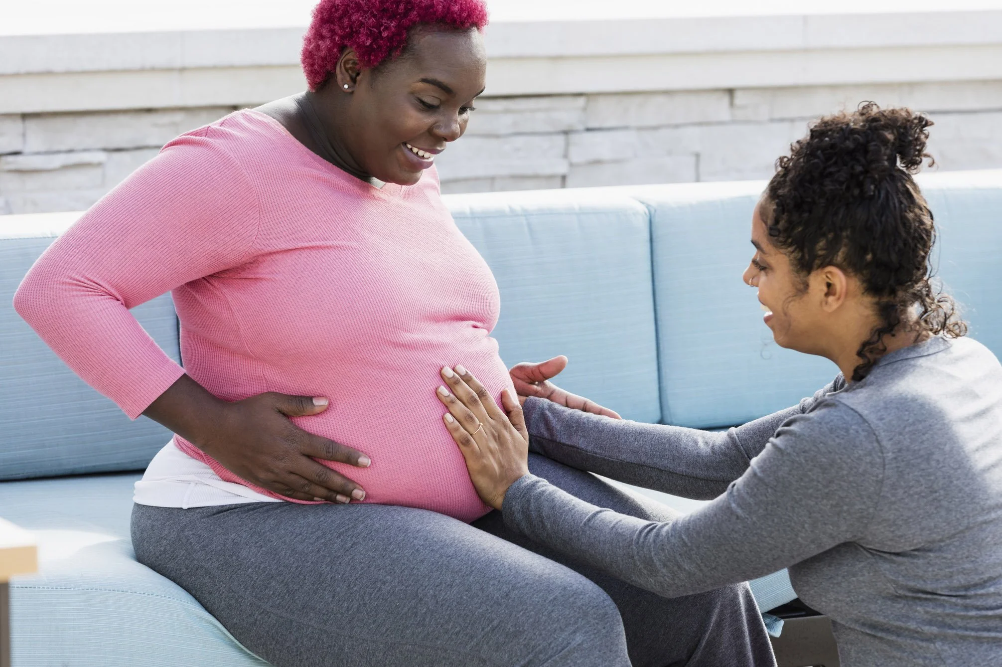 A pregnant Black woman with bright pink hair sitting on an outdoor sofa, smiling down as someone gently places a hand on her baby bump.