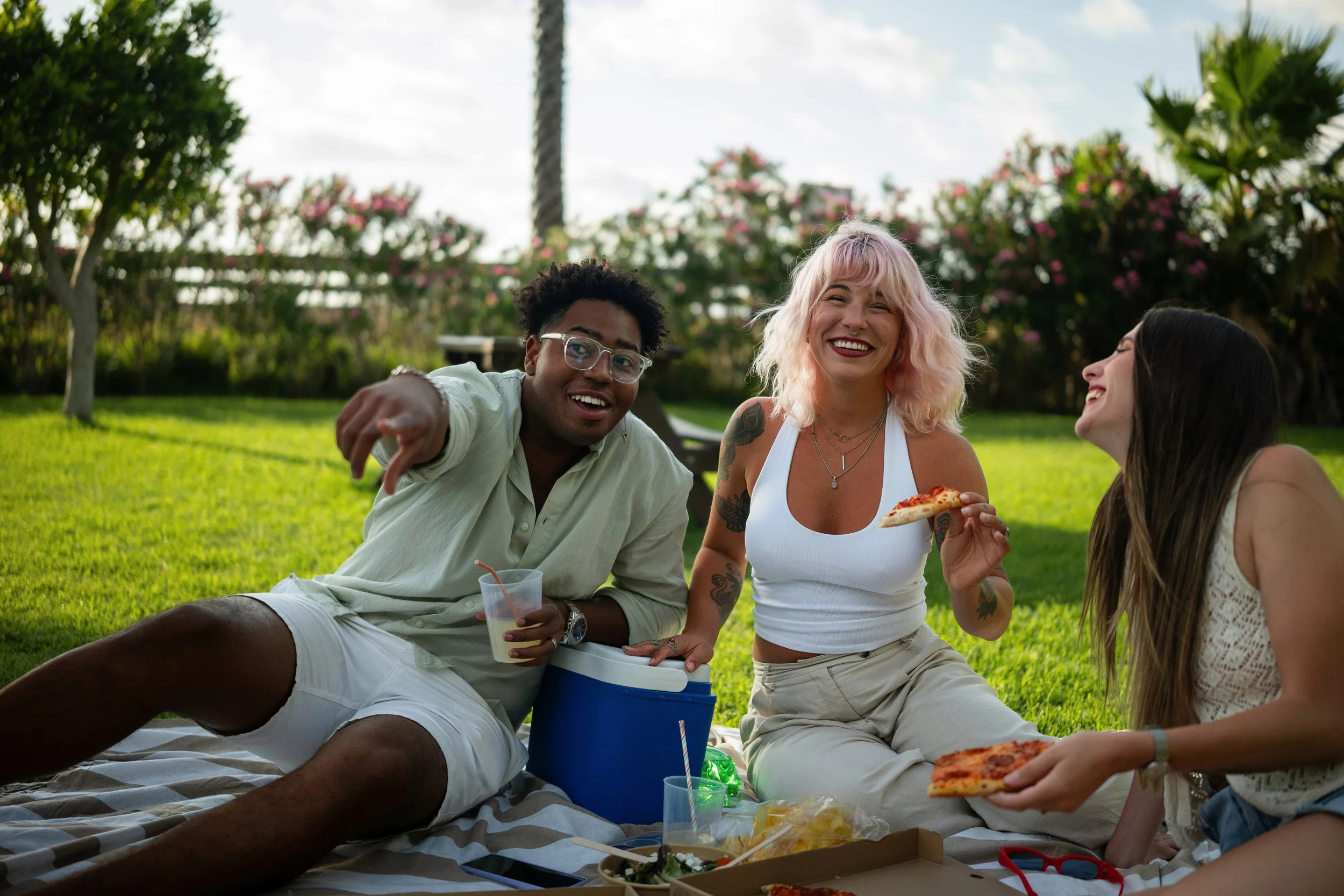 Three young friends laughing and eating pizza together during a sunny outdoor picnic on the grass.