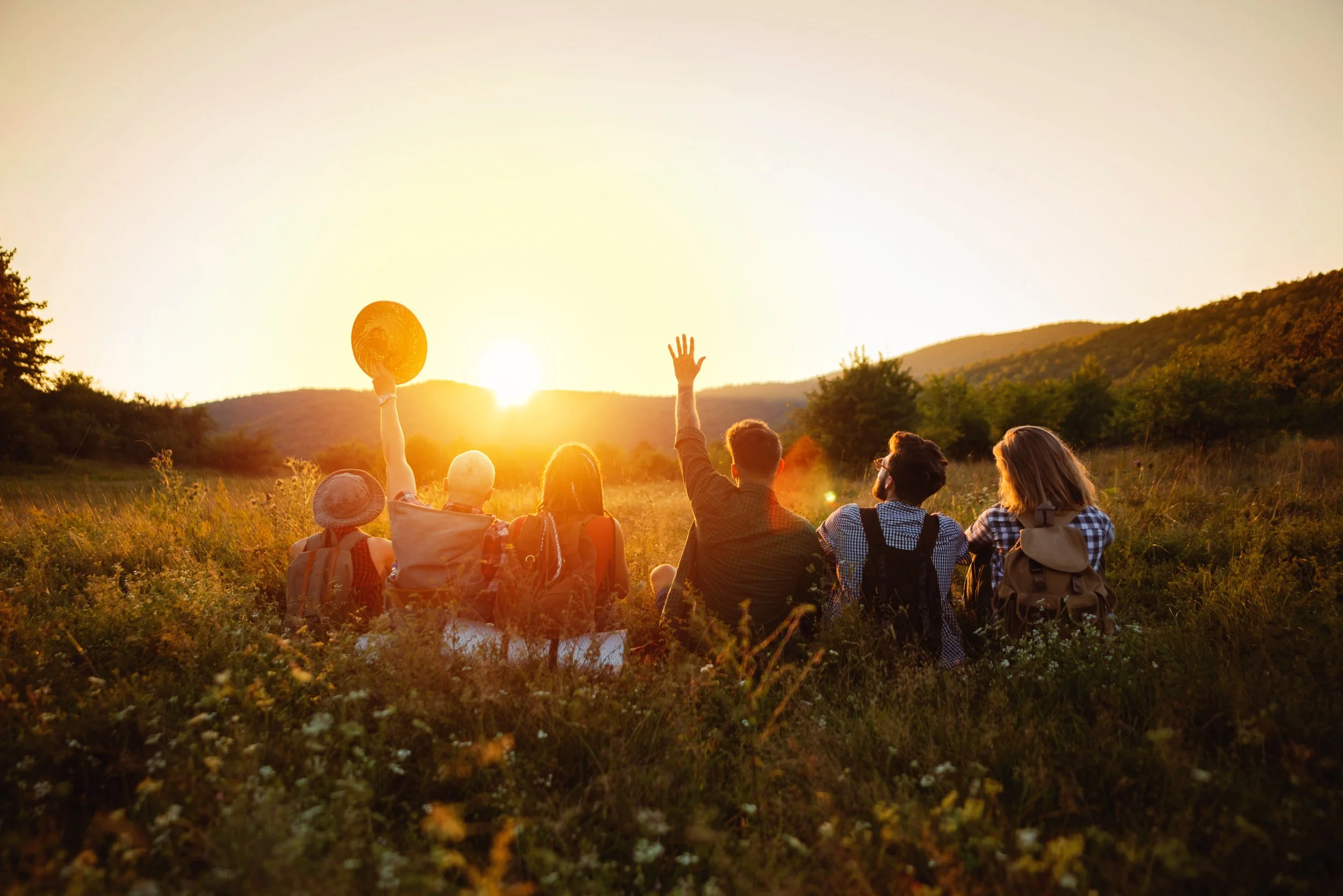Group of young people sitting facing away from camera and raising hands celebrating a sunset in front of them.