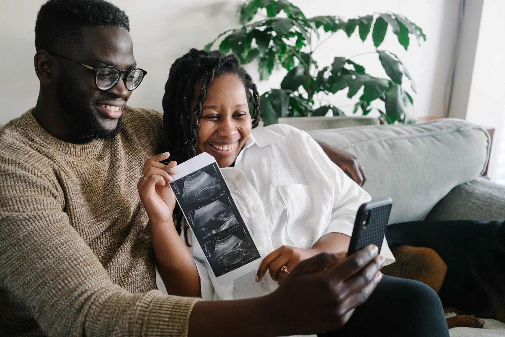 A smiling Black couple sitting on a sofa holding up a strip of baby ultrasound photos for a smartphone video call.