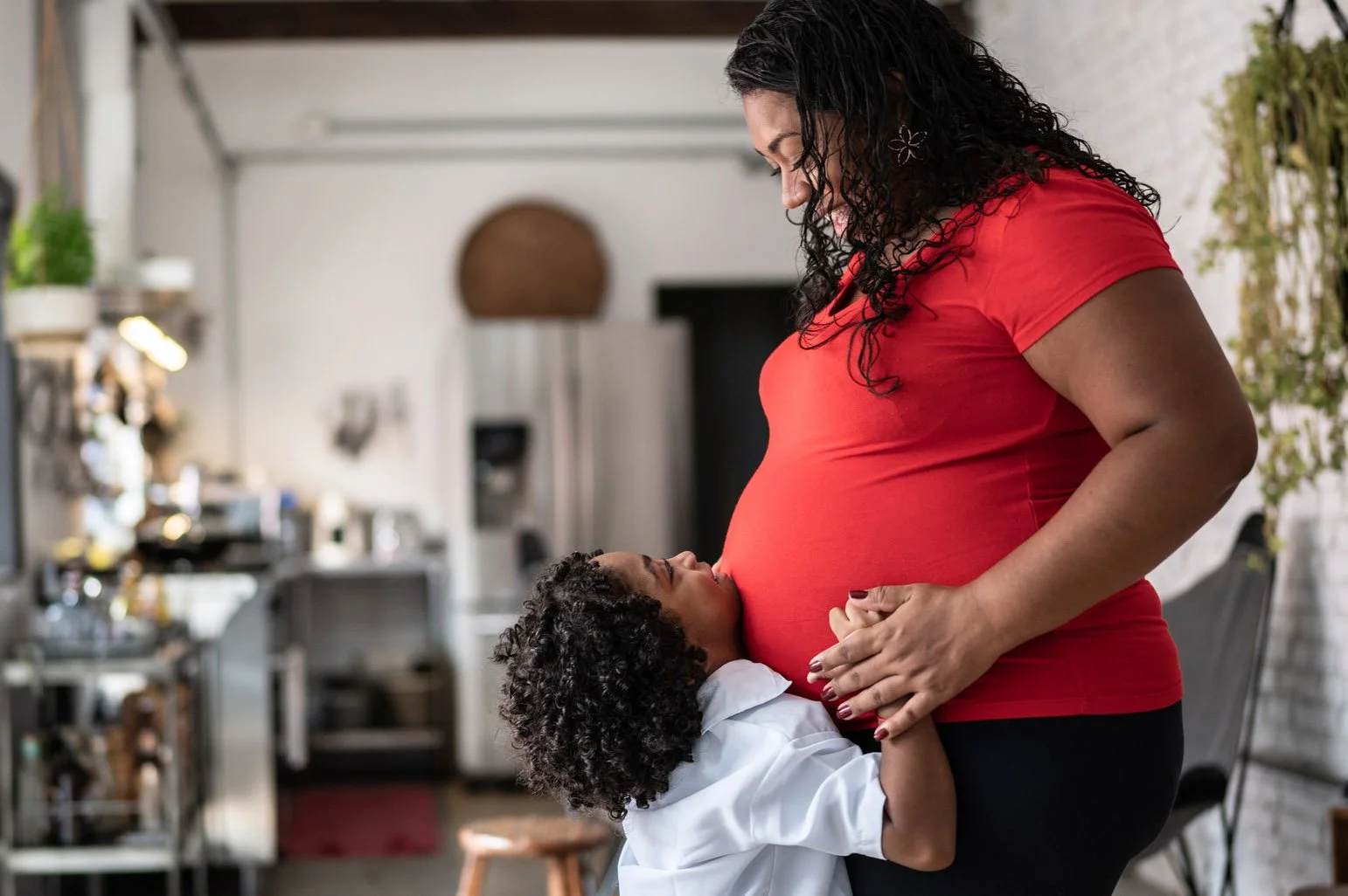 A pregnant mother in a red shirt smiling down at her young child, who leans lovingly against her baby bump while holding her hand.