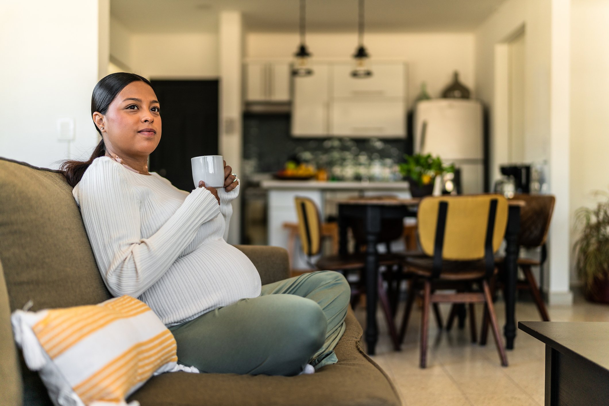 A pregnant woman sitting cross-legged on a living room sofa, holding a white mug with both hands and looking thoughtfully off to the side.