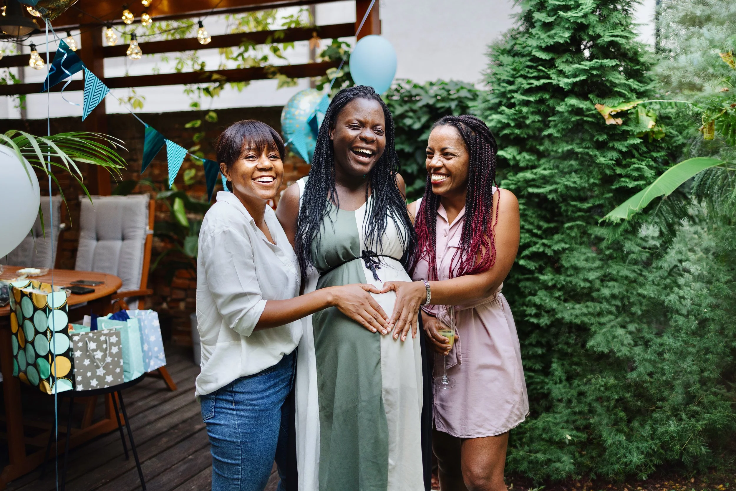 Three Black women laughing outdoors at a baby shower as two of them make a heart shape with their hands over the center woman's pregnant belly.