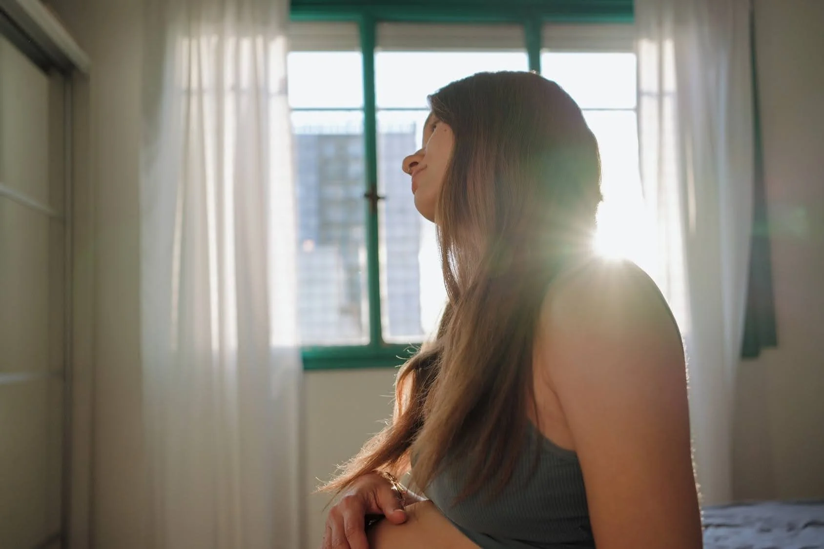 A woman with long brown hair sitting near a window, holding her pregnant stomach in streaming sunlight.