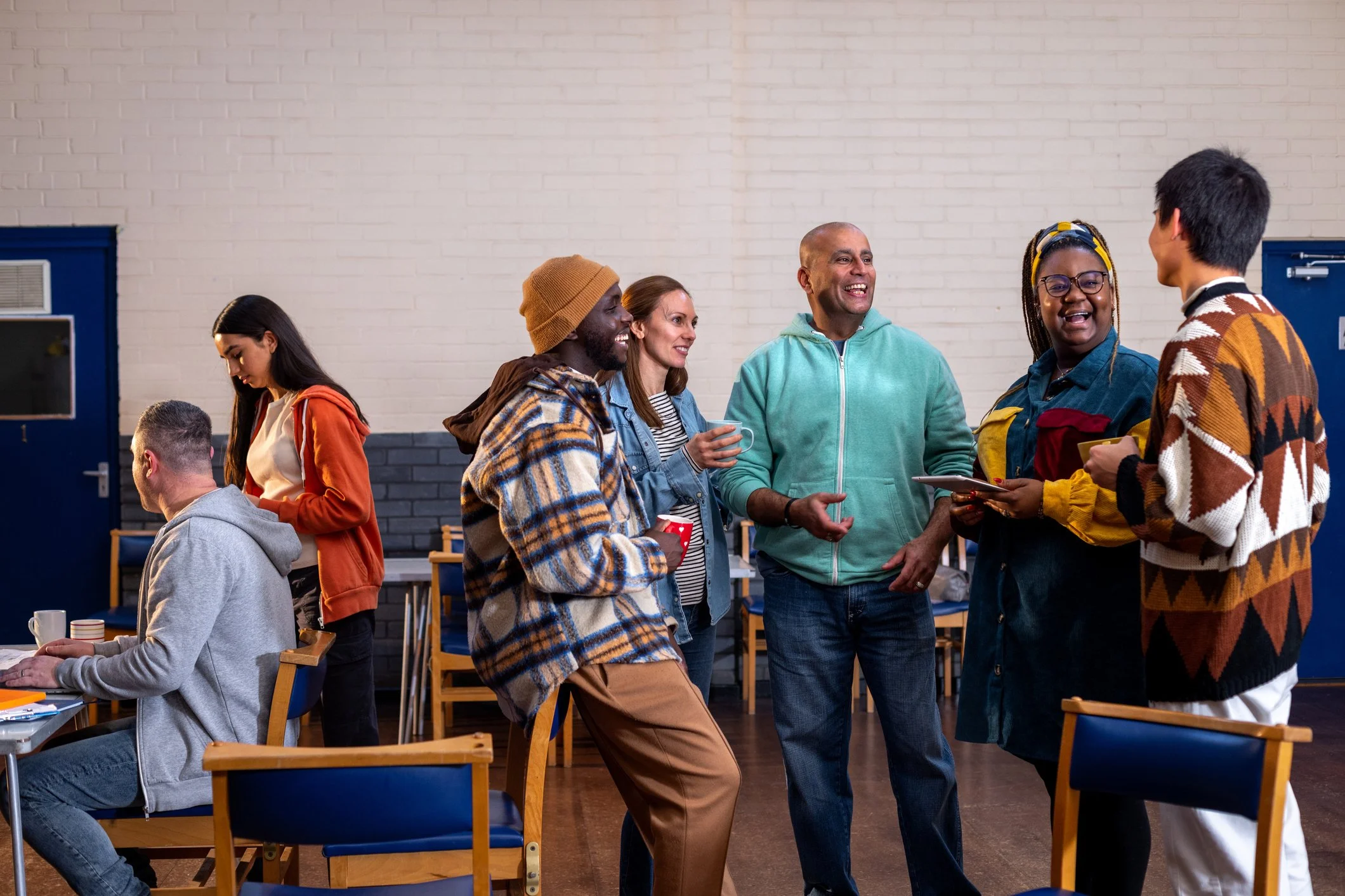 A diverse group of adults chatting, laughing, and holding warm drinks in a community hall or breakroom.
