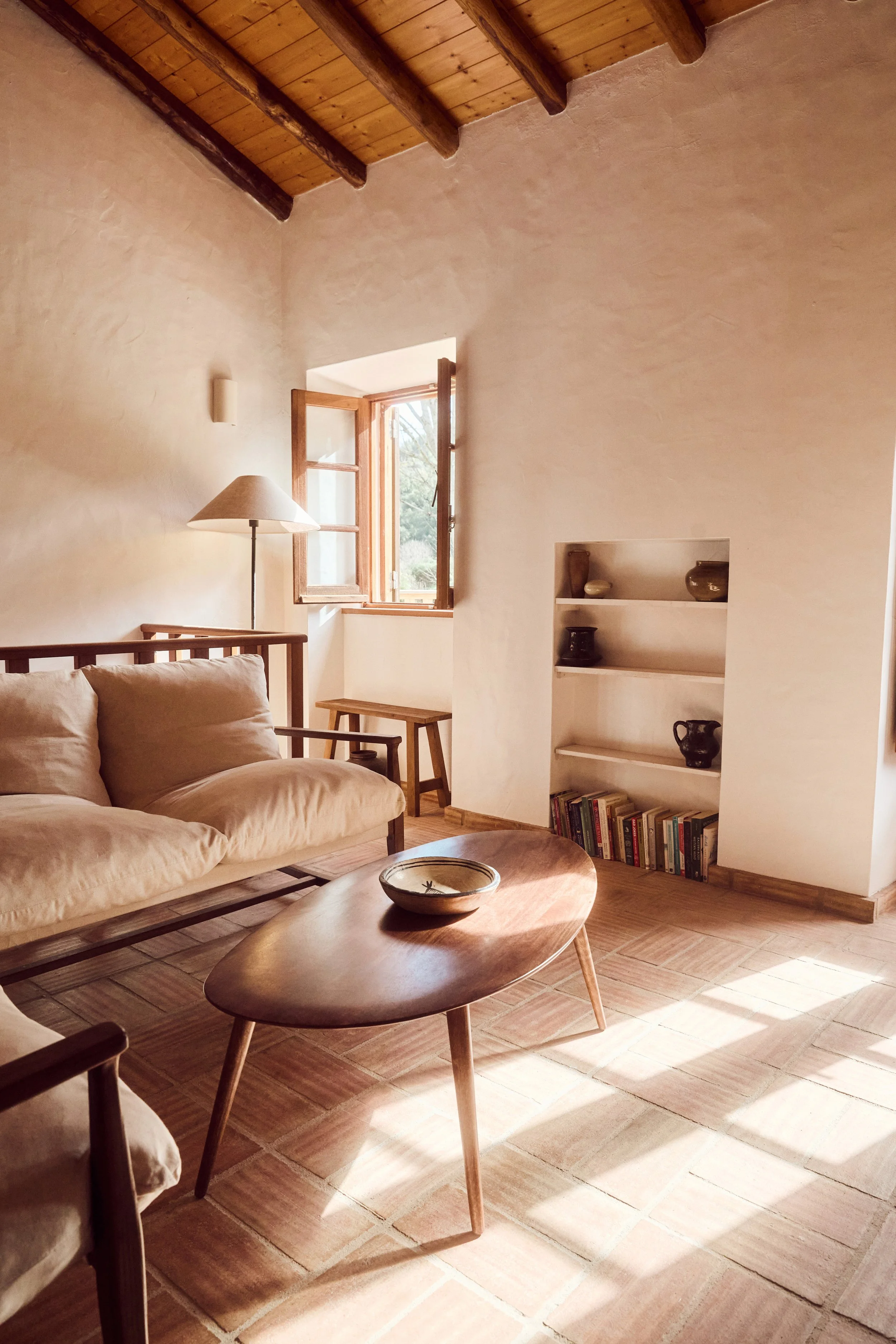 Living room with beige sofa, wooden coffee table, open window, bookshelf, and sunlight on terracotta tile floor in a rustic style with exposed wooden beams.