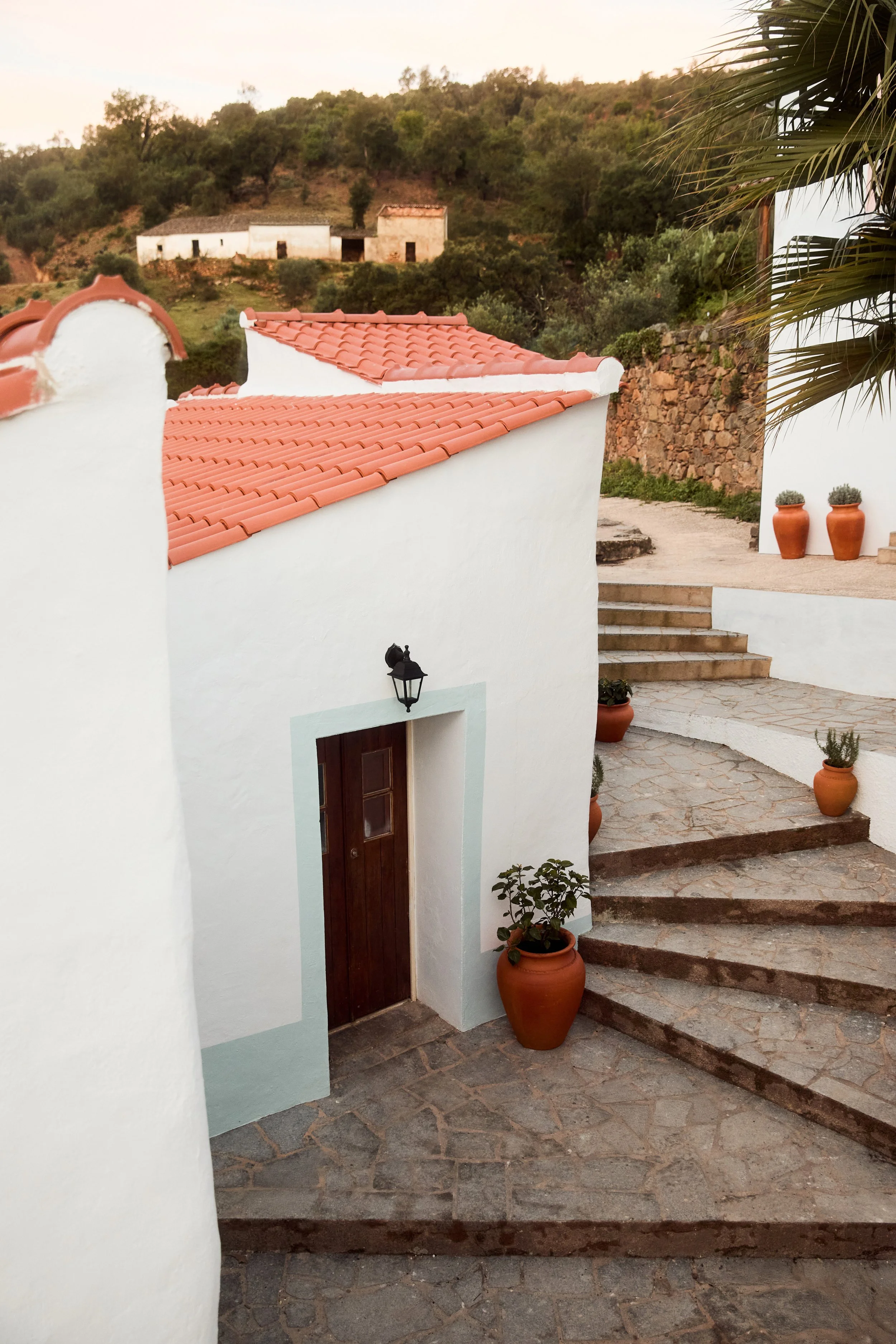 White stucco building with red tile roof, potted plants, stone stairs, and a lantern outside, set against a hillside with trees and additional buildings in the background.