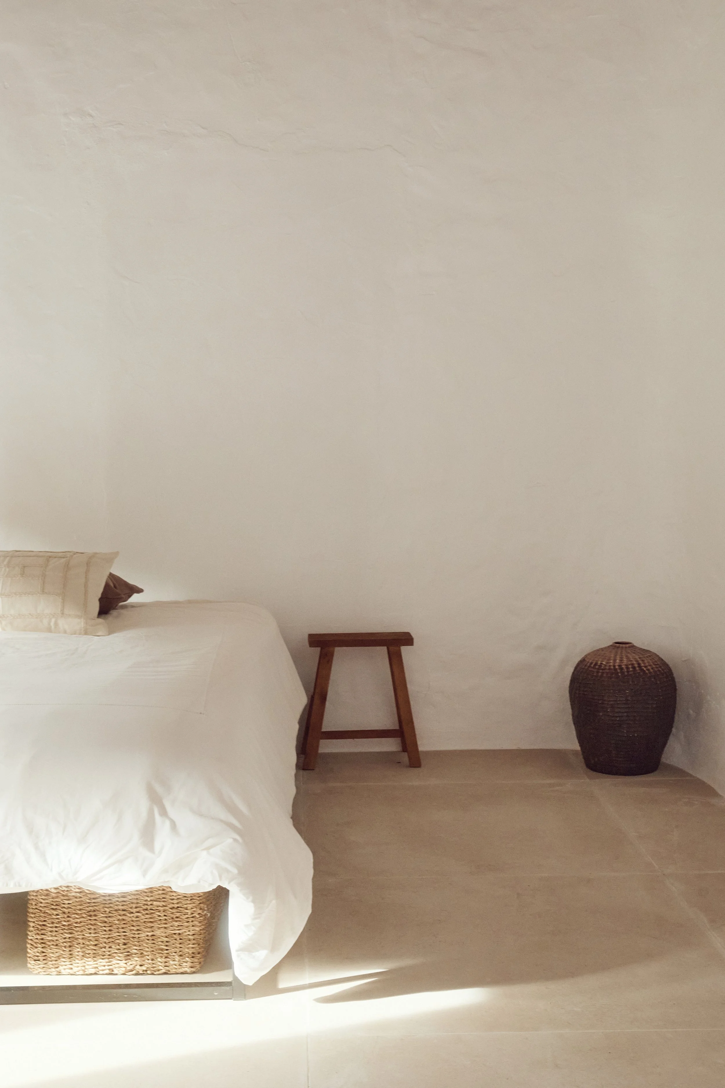 Minimalist bedroom with a bed, a small wooden stool, and a woven basket against a white wall and beige floor.