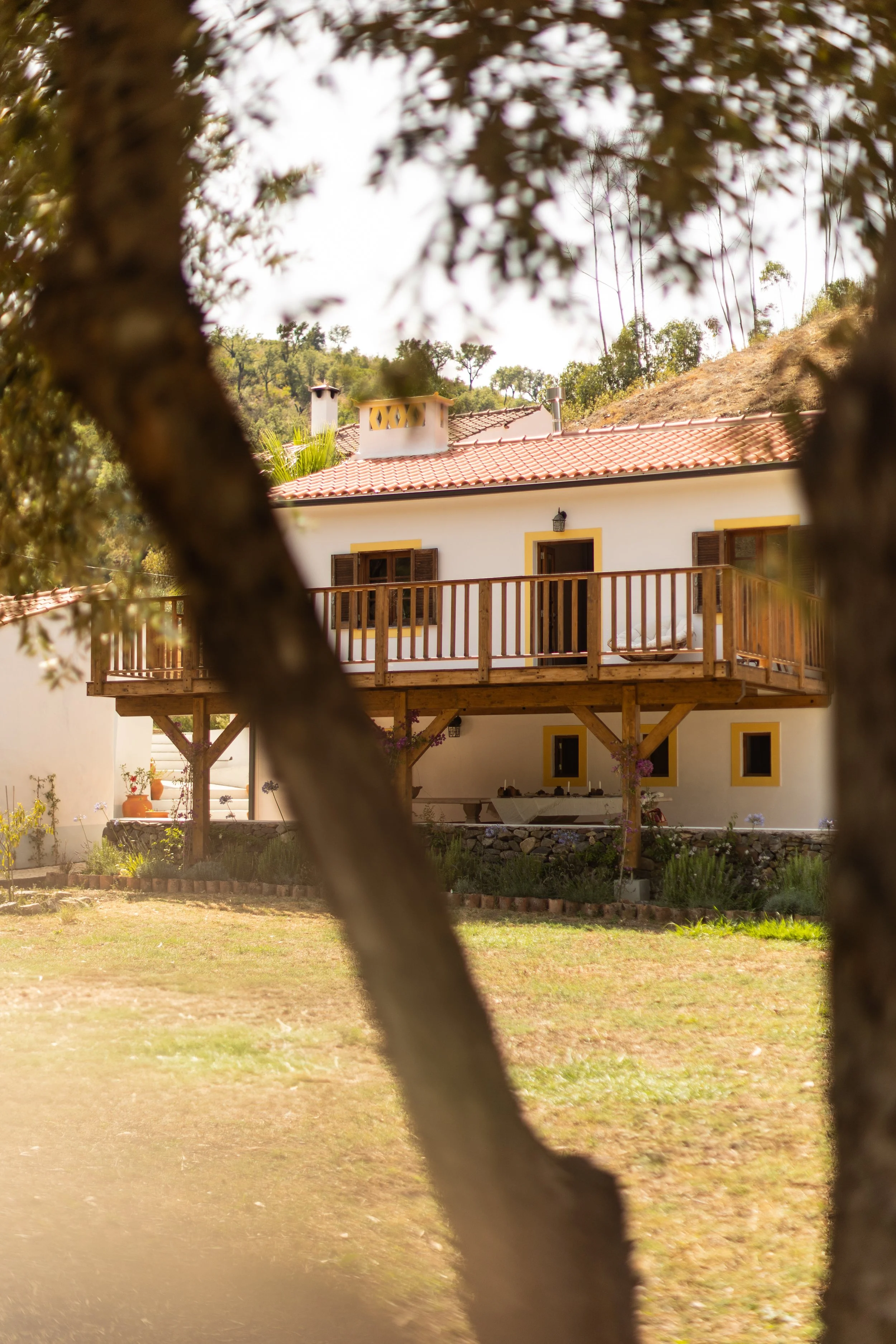 A house with a red-tiled roof, white walls with yellow trim, wooden balcony, and door, viewed through tree branches in the foreground.