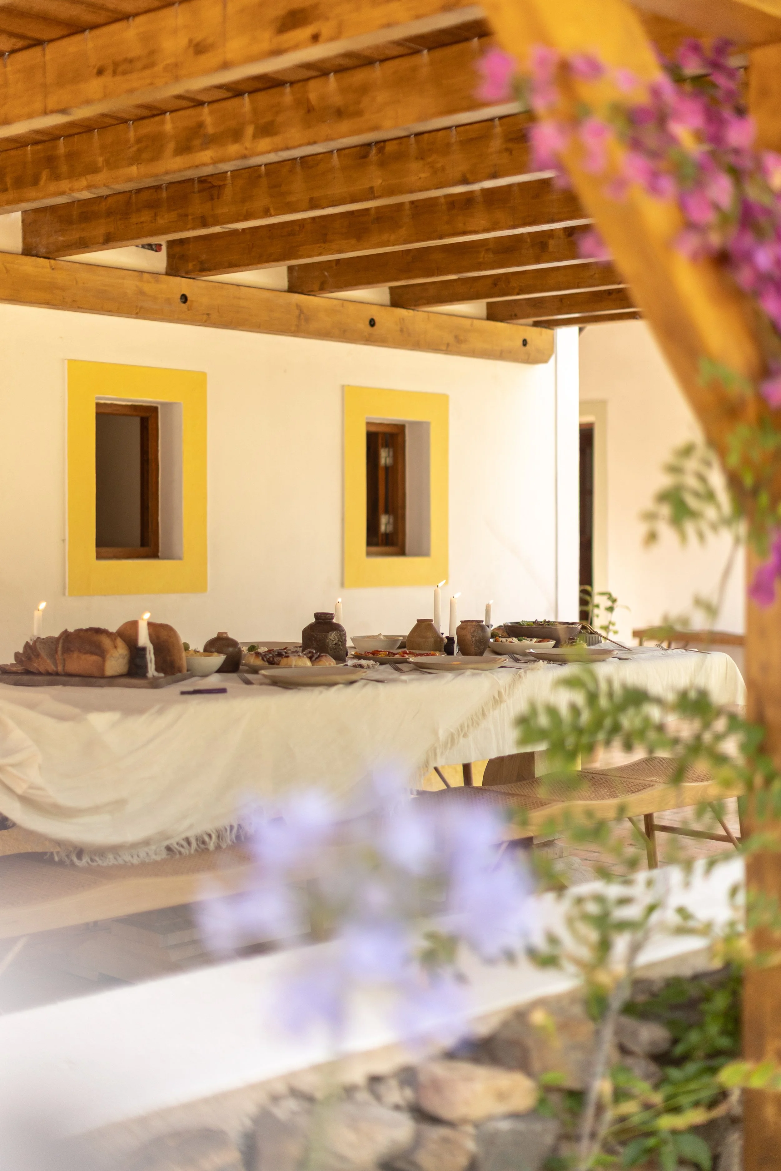 A long dining table with white tablecloth, set with bread, dishes, pottery, and white candles, in a rustic room with wooden ceiling beams, beige walls with yellow-framed windows, and purple flowers in the foreground.