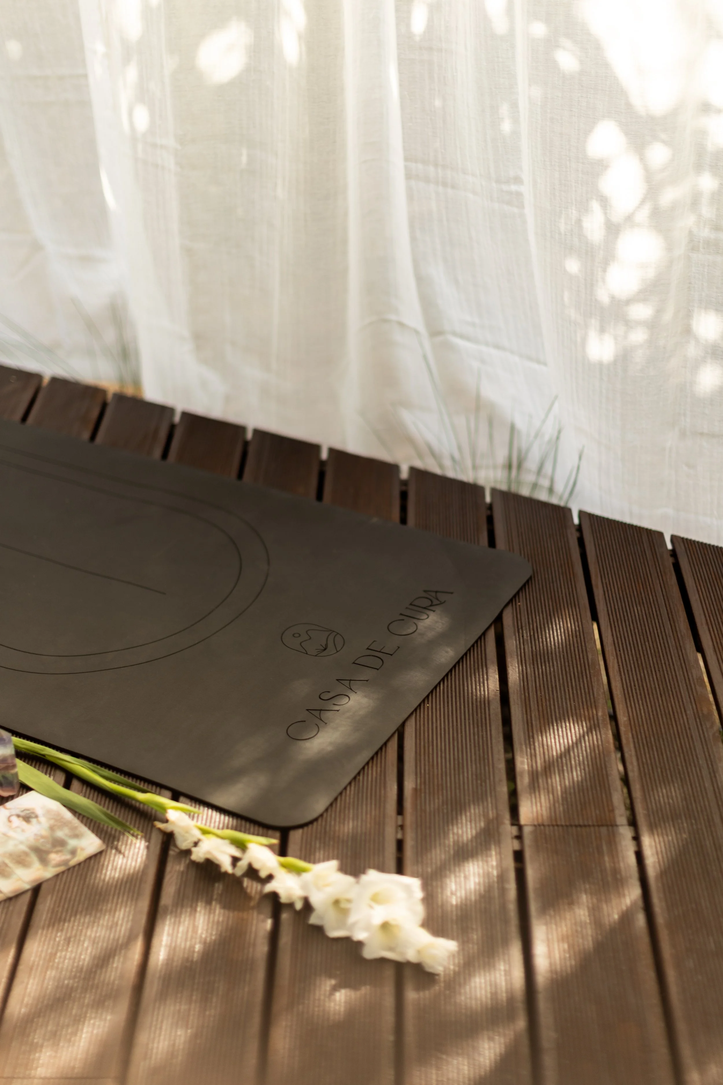 A black induction cooktop with the words "Casa de Cura" and a small logo, placed on a wooden table next to a white flower.