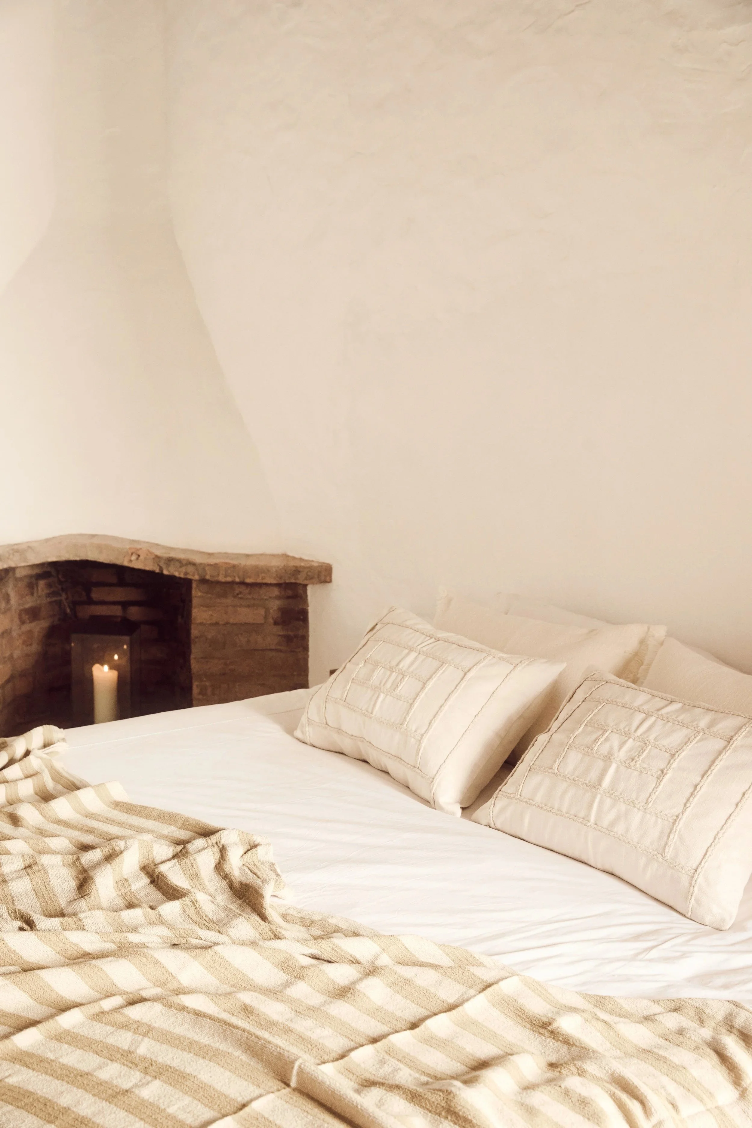 A cozy bedroom with a white bed, cream pillows, a beige striped blanket, a brick fireplace with a candle inside, and white walls.