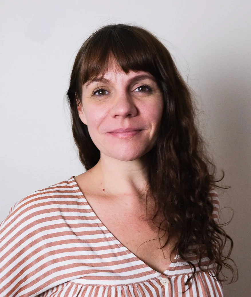 A woman with long, wavy brown hair wearing a striped top, smiling softly at the camera against a plain white background.