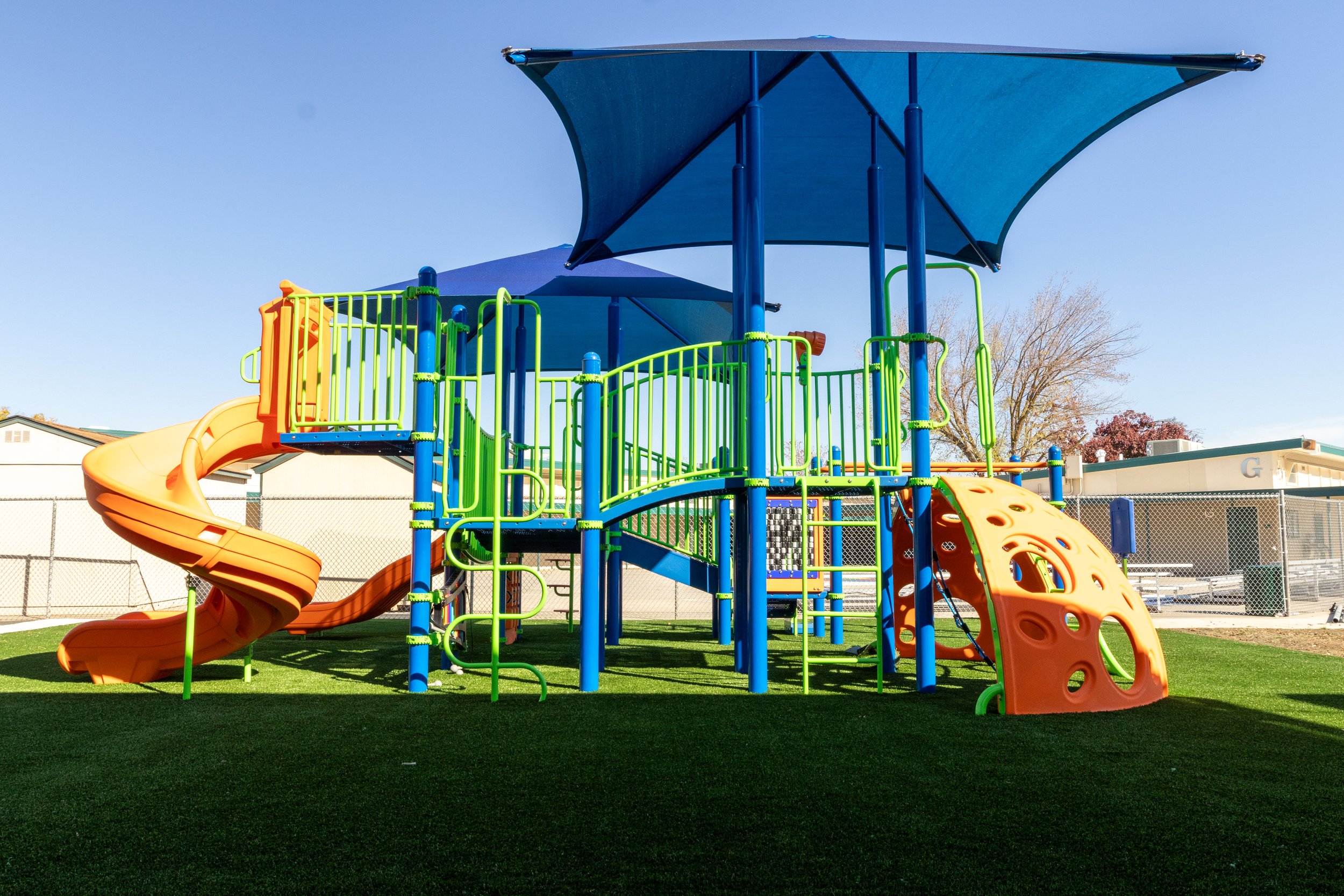 Colorful playground equipment with slides, climbing structures, and blue shade canopies on artificial grass.