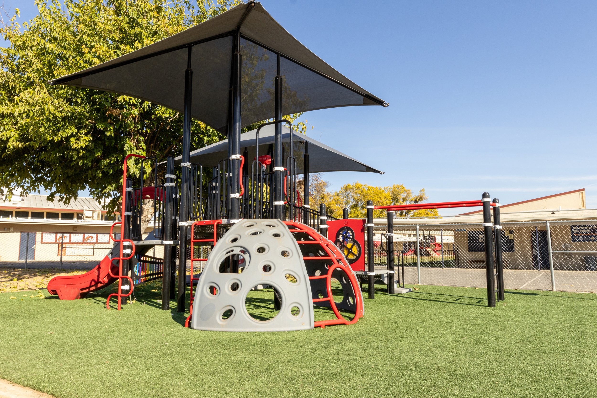 Modern playground with slides, climbing structures, and shade canopies on artificial grass surface, surrounded by a fence and trees.