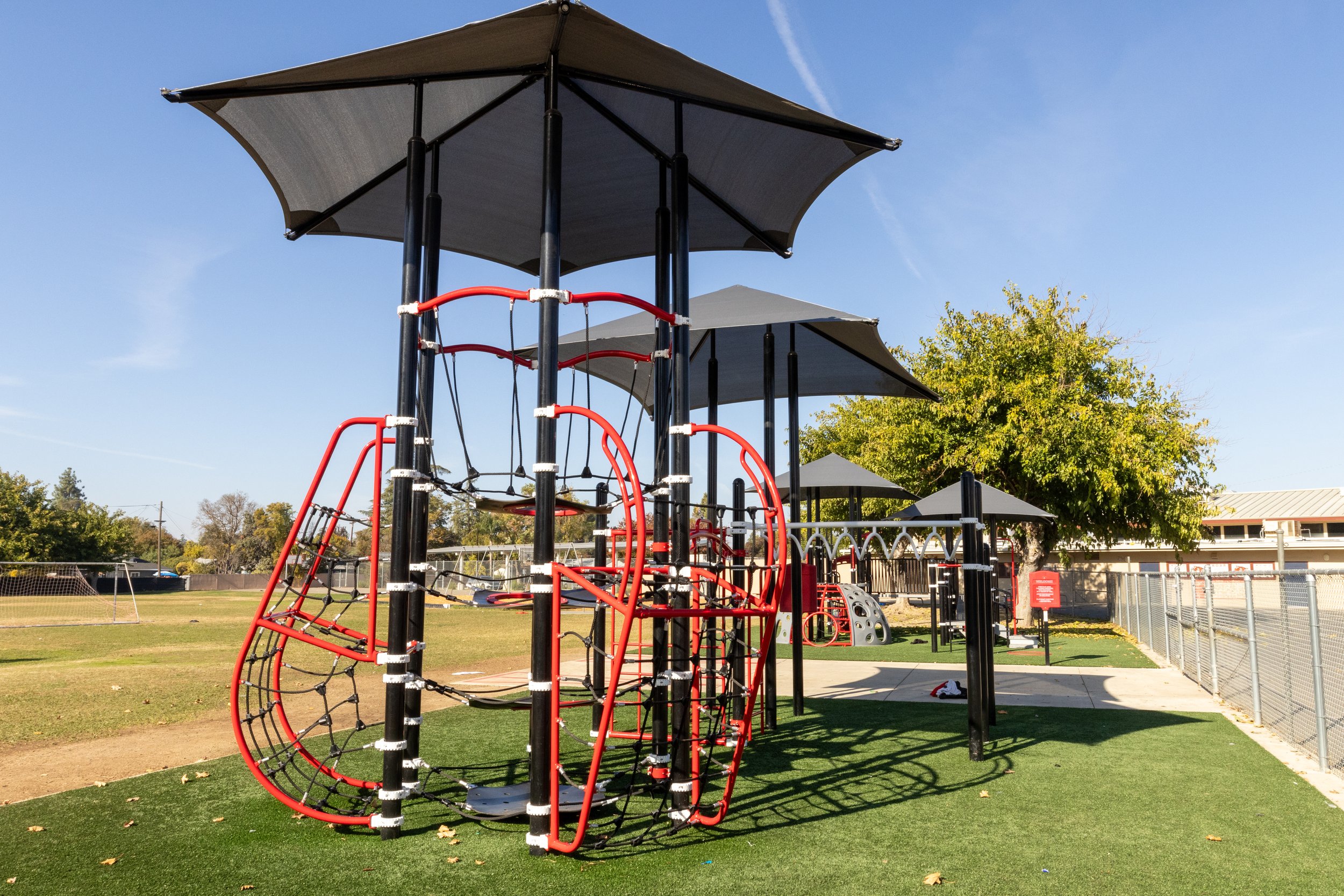 Outdoor playground with modern climbing structures, featuring black and red frames, under shade canopies. The playground is set on artificial turf surrounded by a fence, with trees and a building in the background.