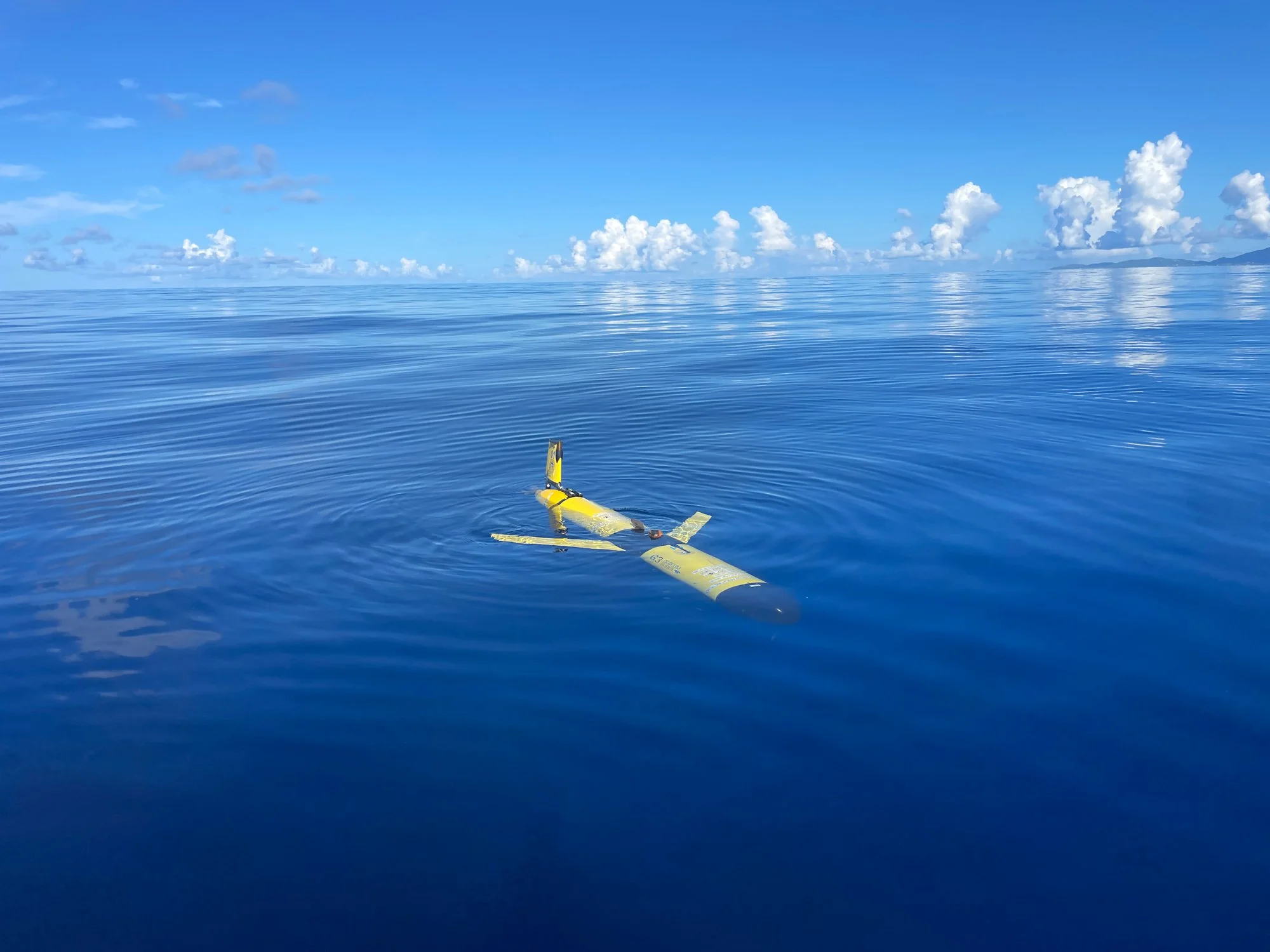 An ocean glider on the surface of open water off St. Thomas, with a research vessel behind it. Credit: UVI Glider Lab
