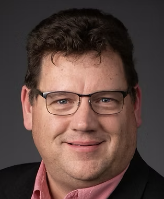 Close-up portrait of a man with glasses, smiling, wearing a dark suit jacket and a pink shirt, against a dark background.