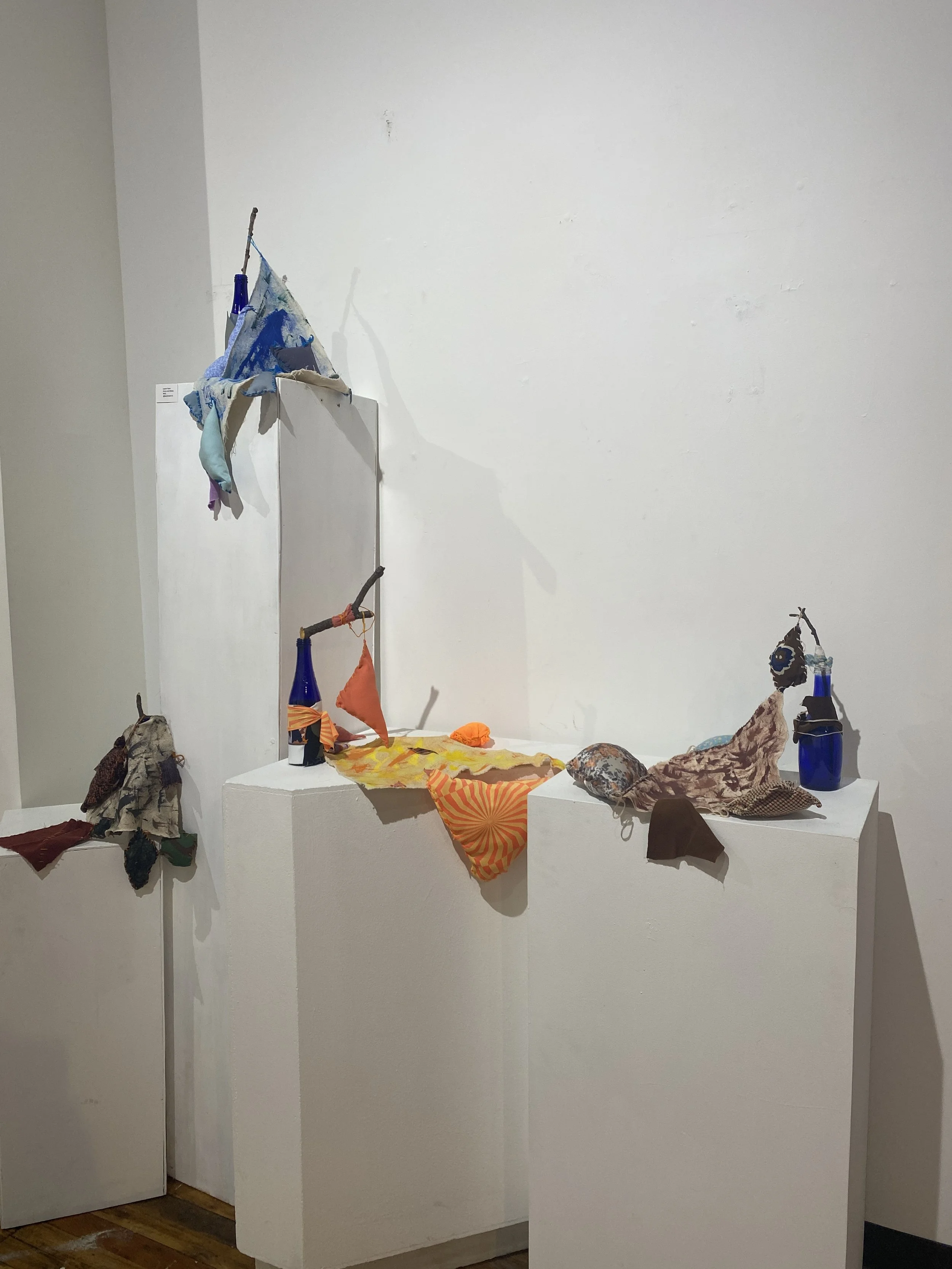 Above and Below* (Series), 2022. Acrylic paint, recycled fabric filled with synthetic fiber to resemble a plush toy, and embroidery floss on canvas installed using found twigs in repurposed glass bottles; Blue triangle: 8" x 26.5" x 5", Orange triang