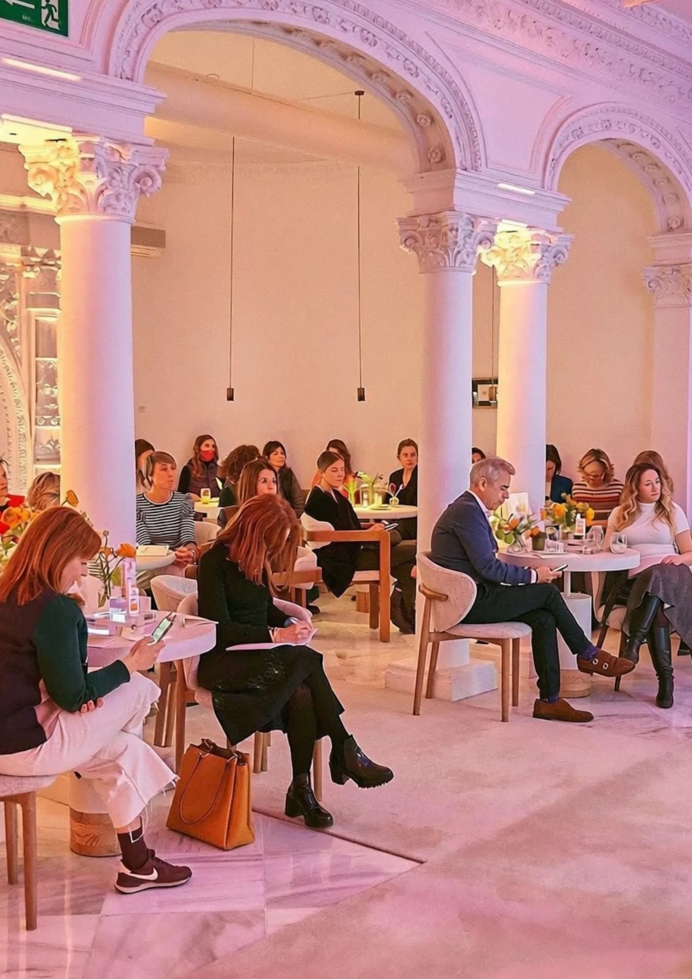 Una sala elegante con columnas y arcos decorativos en tonos rosados, donde varias personas están sentadas en mesas redondas, algunas usando teléfonos móviles y otras observando, en un ambiente iluminado con luces cálidas y decoraciones florales.