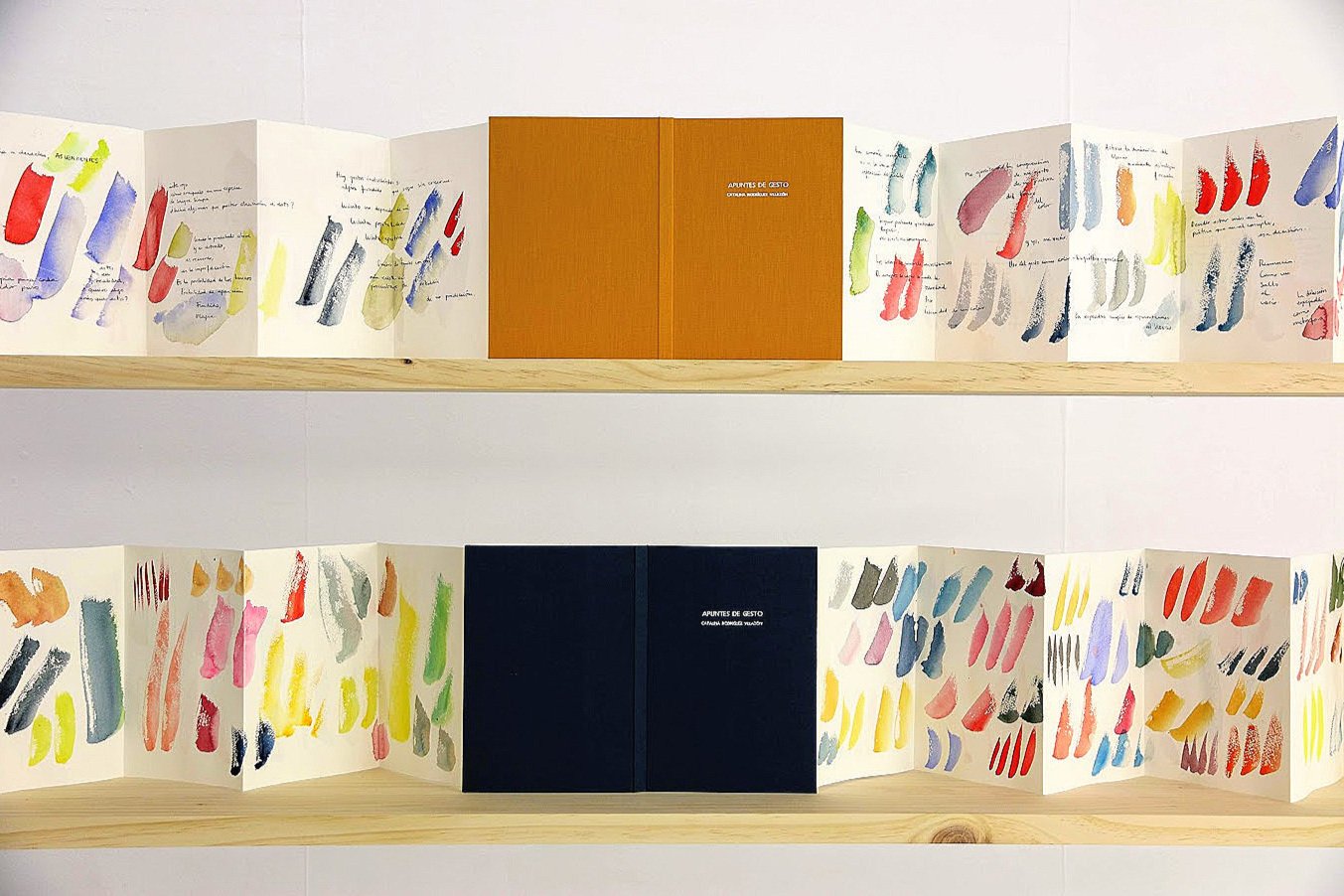 Dos estanterías con libros abiertos mostrando páginas con patrones de manchas de acuarela en varios colores y letras manuscritas, con libros centrales de portada naranja y azul.