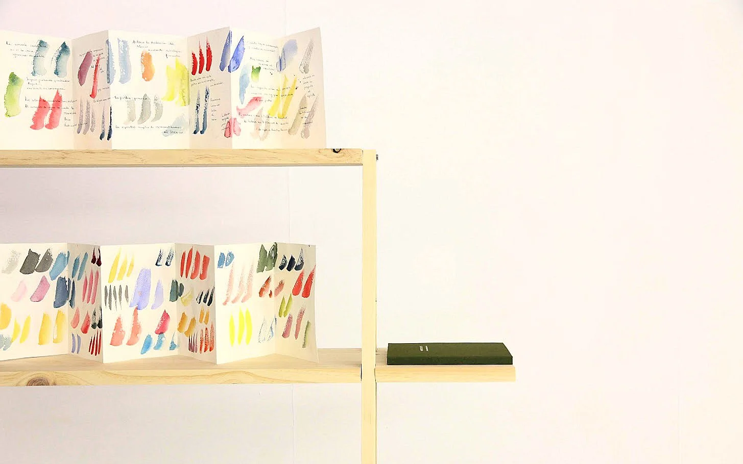 Muestras de diferentes patrones y colores de pintura en un folleto desplegado en una estantería de madera, ubicada en una pared blanca, con un libro negro sobre una repisa adicional.