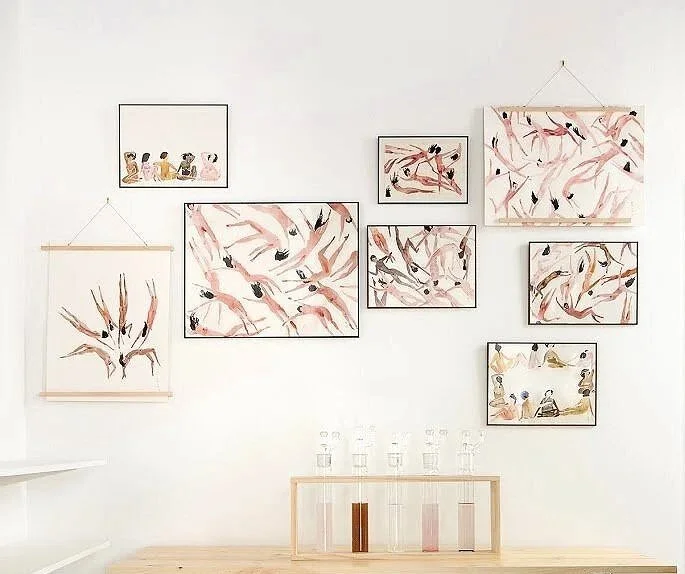 Conjunto de cuadros con pintura abstracta y figuras humanas en colores suaves, en una pared blanca decorada con un mueble de madera y pequeños frascos.