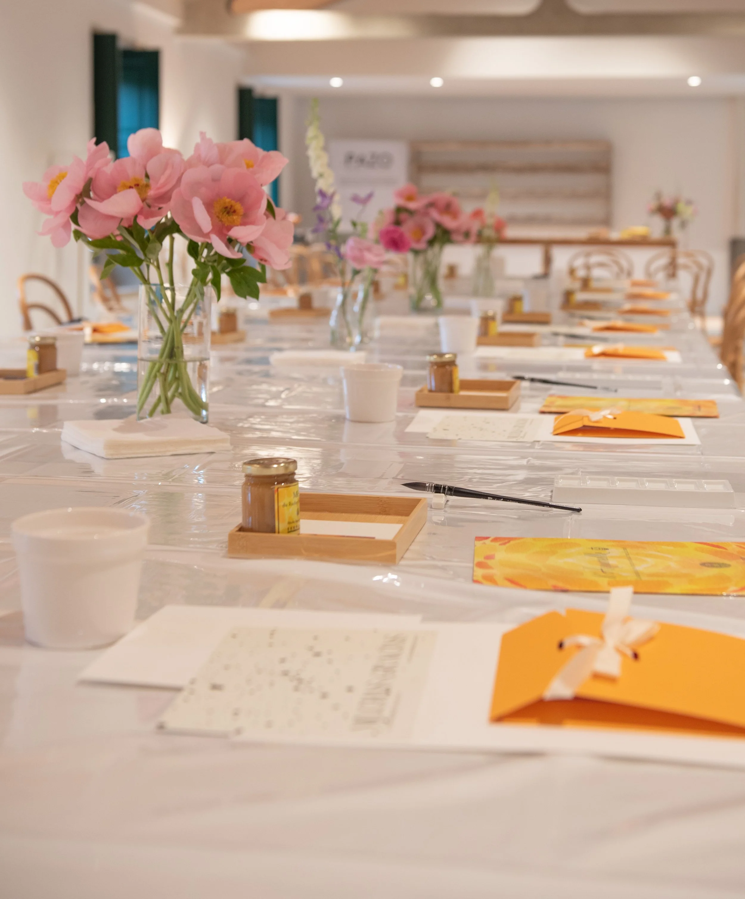 Mesa larga con varias flores en jarrones, papeles, tazas, y pequeños frascos, en una sala bien iluminada y decorada con cortinas azules.