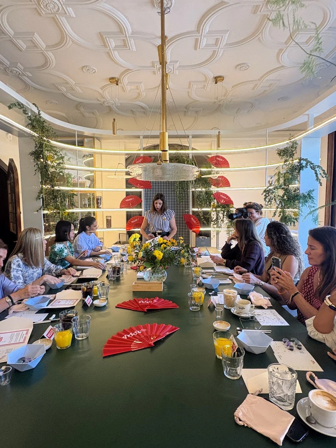 Reunión con varias personas sentadas a una mesa rectangular en un comedor elegante con decoraciones verdes y rojas, en el centro hay un jarrón con flores amarillas, y un cuadro en la pared del fondo. Una mujer de pie en el centro parece estar liderando la reunión, mientras otra con cámara captura el evento, y varias personas están usando celulares o leyendo.
