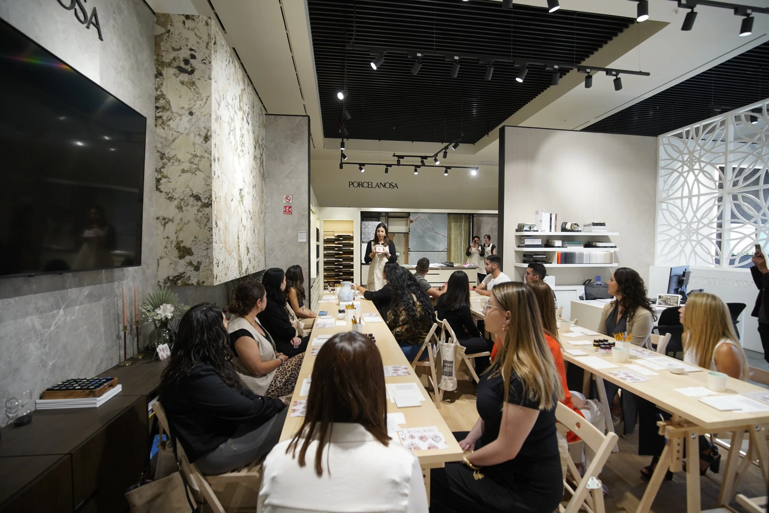 Imagen de una tienda o sala de exhibición de porcelanas y superficies de mármol, con varias personas sentadas alrededor de una mesa y una mujer de pie explicando o presentando, en un ambiente moderno y bien iluminado.