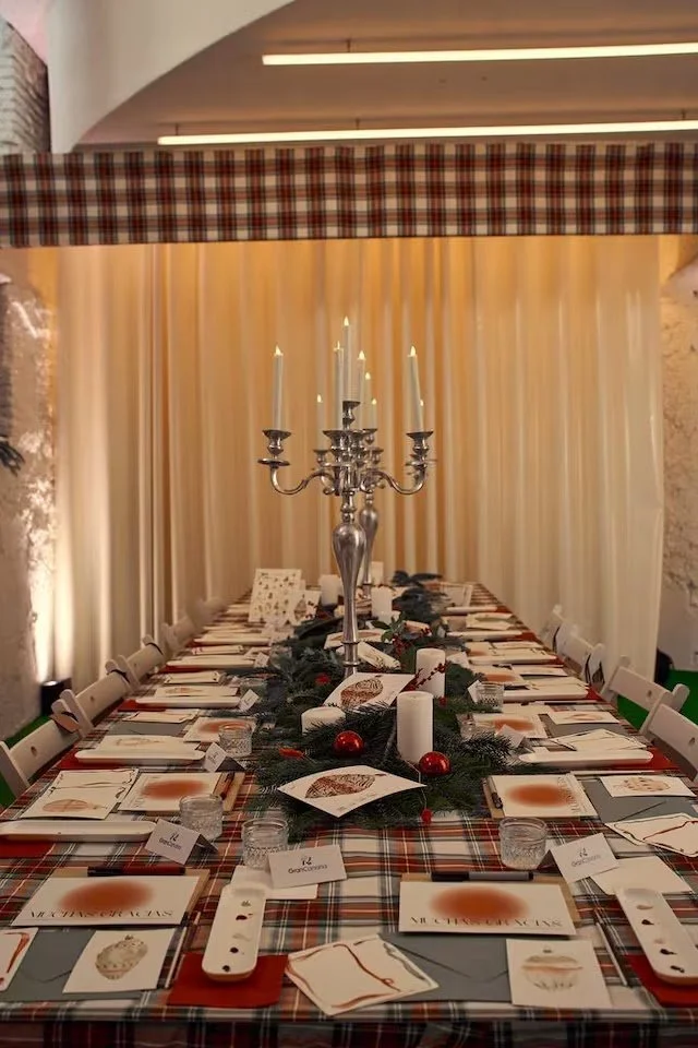 Mesa decorada para una celebración, con candelabro central, velas, adornos navideños, platos, copas y servilletas en la mesa, en un ambiente con cortinas y paredes de piedra.