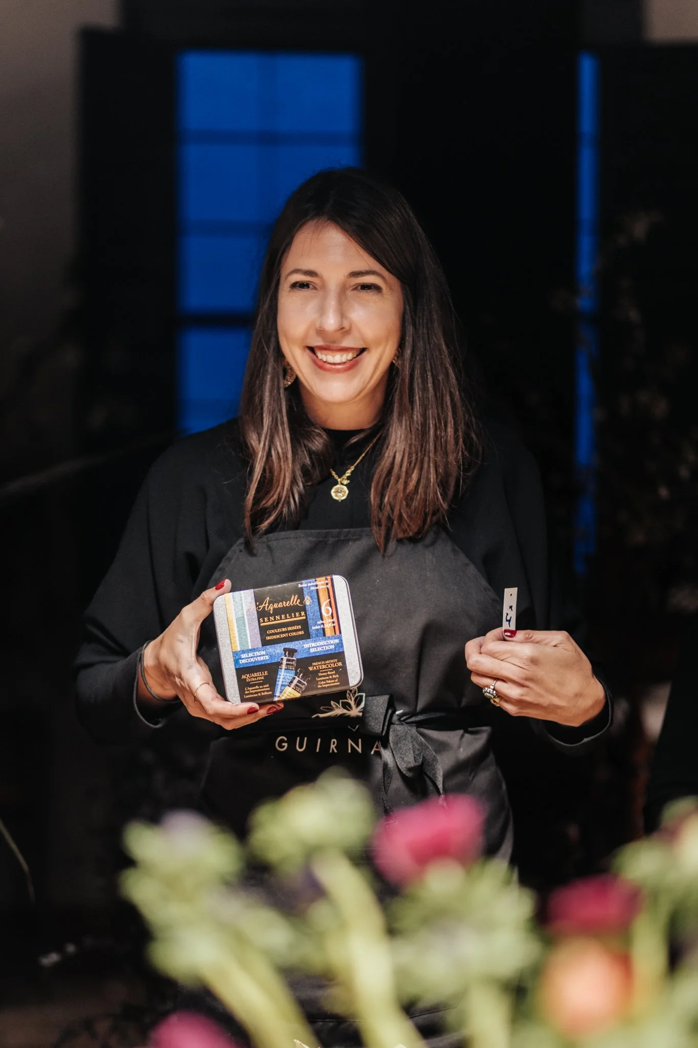 Mujer sonriendo, sosteniendo un kit de acuarelas para pintura y un pincel, en un entorno interior con ventanas azules de fondo. Tiene cabello castaño, lleva un collar dorado y ropa oscura.