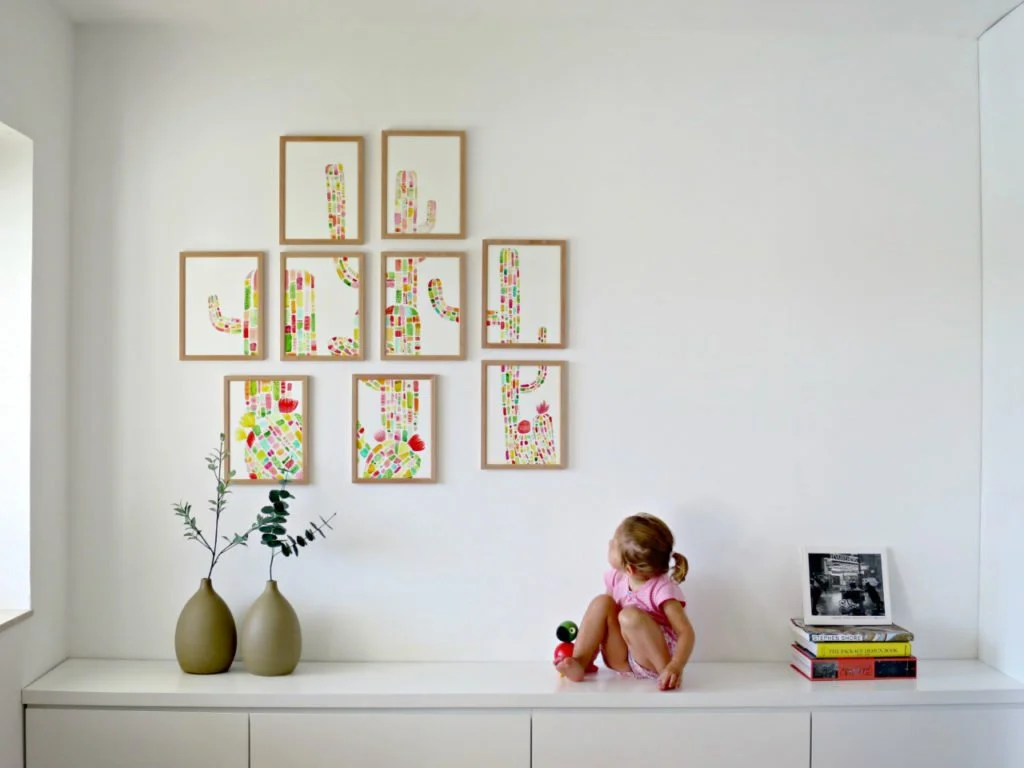 Niña sentada en un mueble blanco, con una pelota de juguete y libros apilados, en una habitación con paredes blancas y decoraciones coloridas.